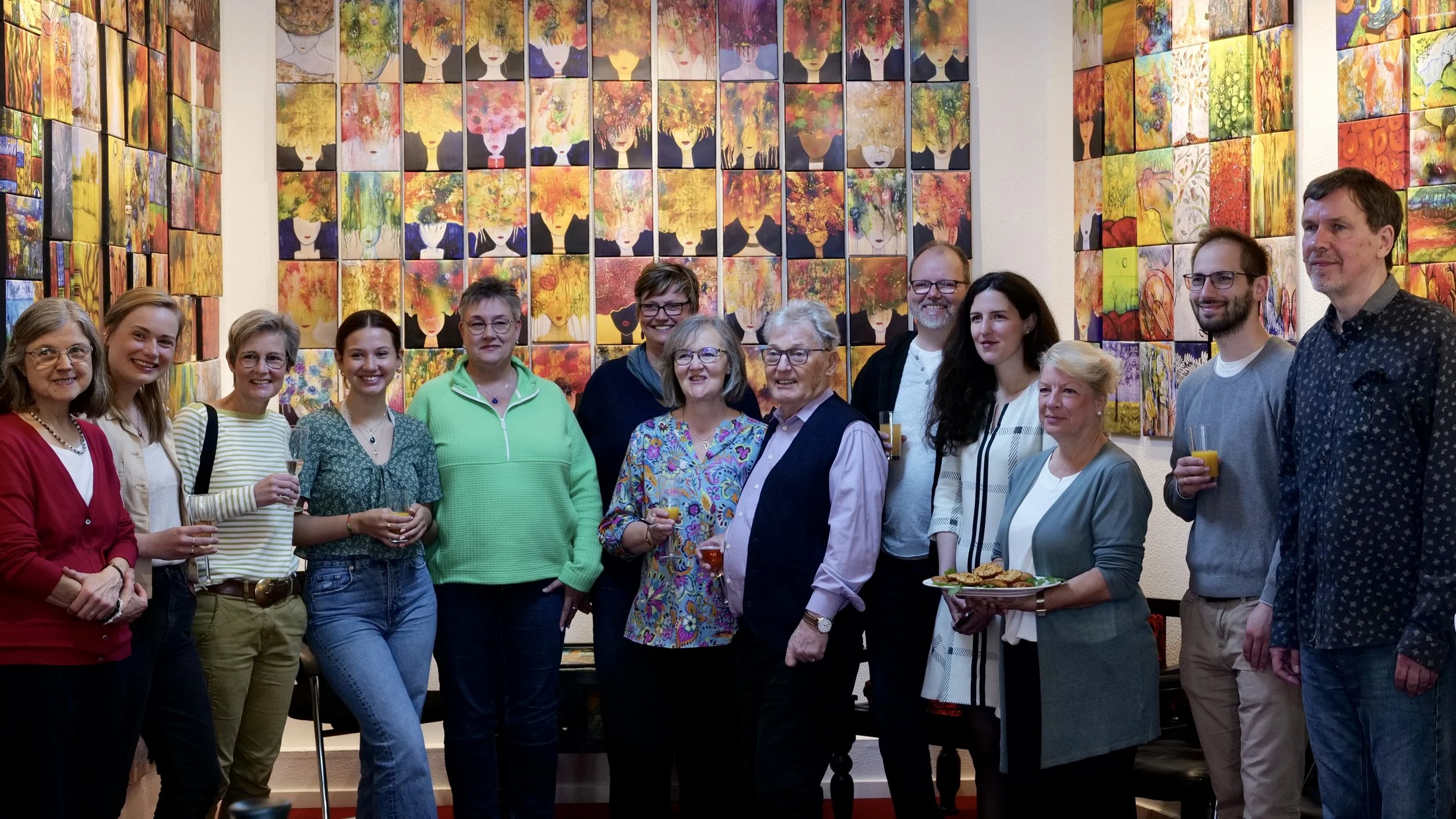 Gruppe von Menschen bei einer Feier in einem Raum mit bunten Gemälden an der Wand, einige halten Getränke und Teller mit Essen.