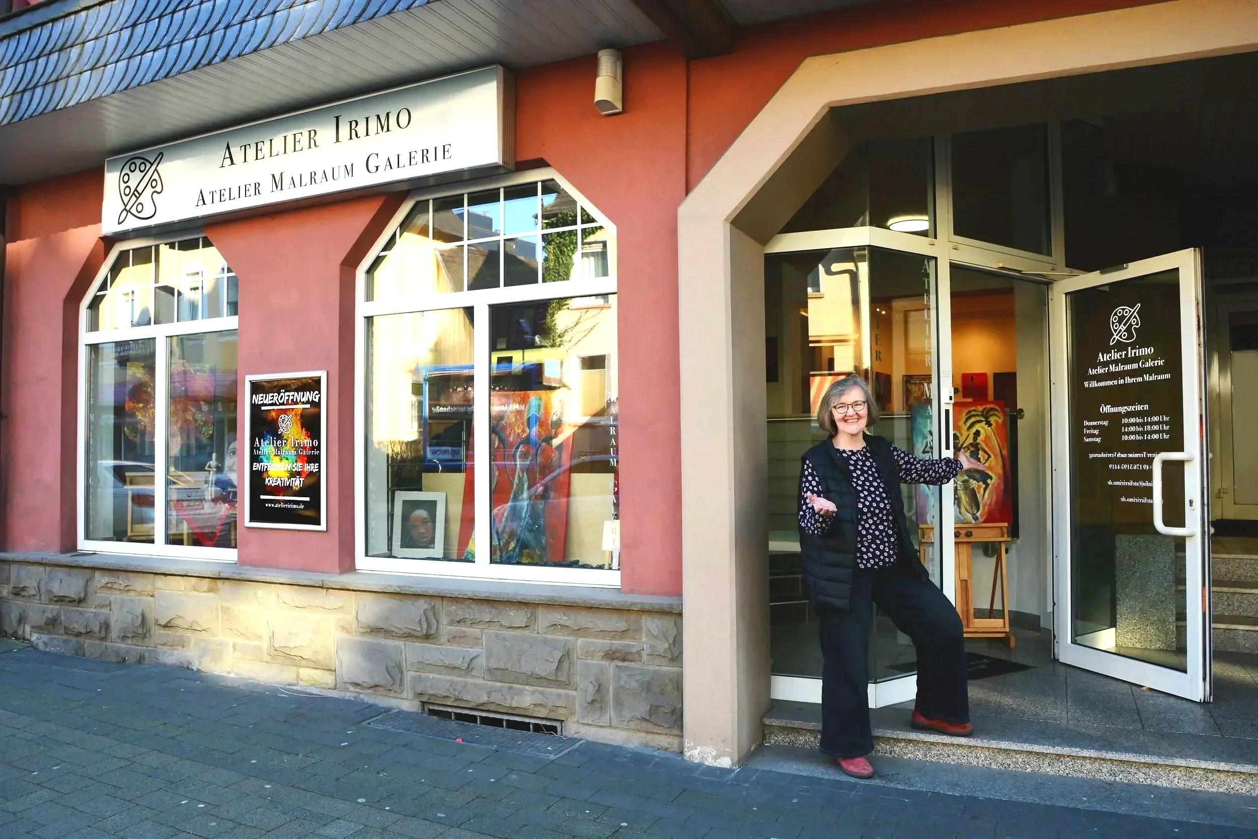 Frau in der Türe eines Kunst- und Galerieraums, die freundlich lächelnd und mit offenen Armen begrüßt.