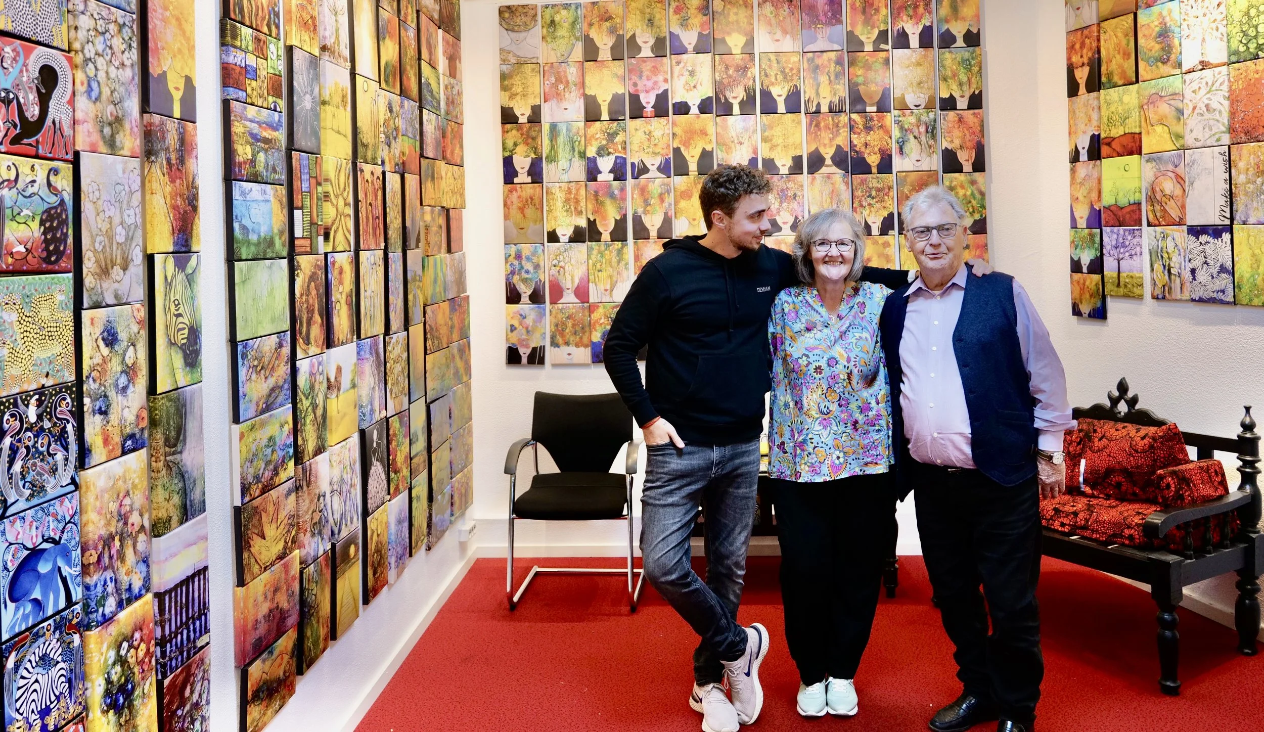 Drei Menschen stehen vor einer Wand aus bunten, abstrakten Kunstwerken in einer Galerie. Es sind eine junge Frau, eine ältere Frau und ein älterer Mann, die alle lächelnd in die Kamera schauen.