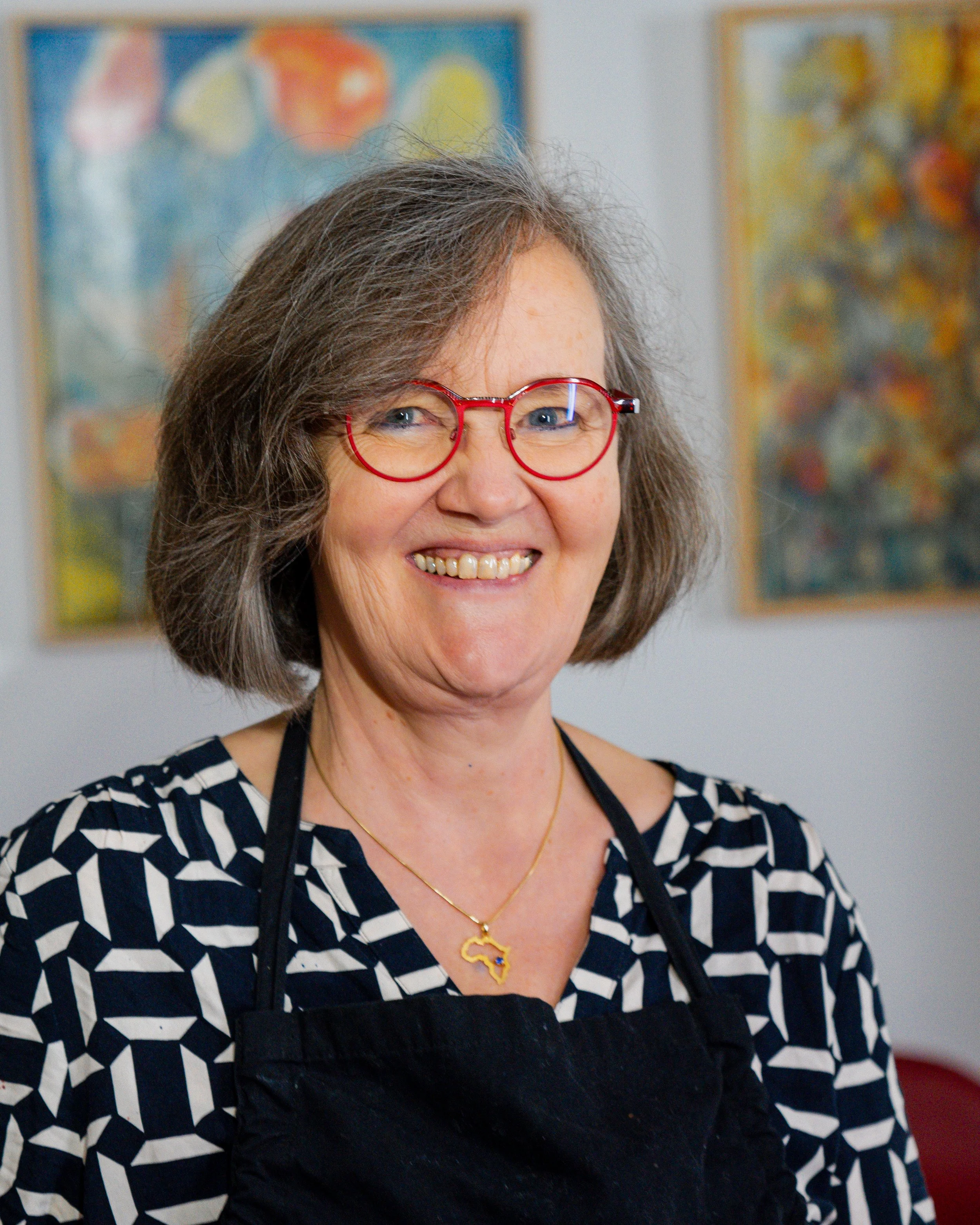 Lächelnde Frau mit grauem Haar, roter Brille, schwarzem Latz, schwarz-weiß gestreiftem Hemd und einer Halskette mit einem Südamerika- und ein Afrique-Schild. Im Hintergrund sind bunte Kunstwerke an der Wand zu sehen.