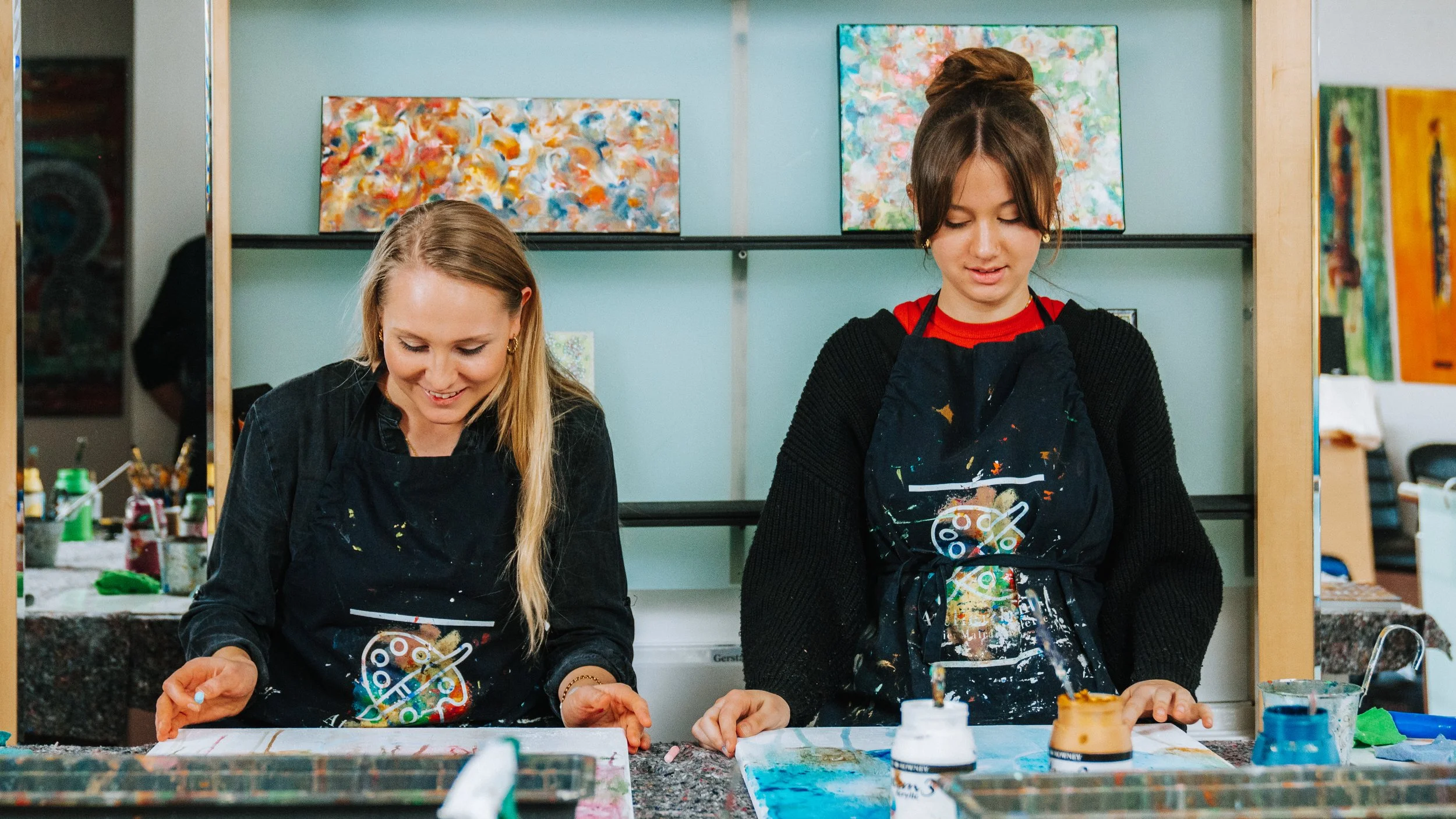 Zwei junge Frauen beim Malen in einem Kunststudio, beide tragen schwarze Schürzen mit bunten Farbspritzern, und stehen vor Tischen mit Farben und Leinwänden.