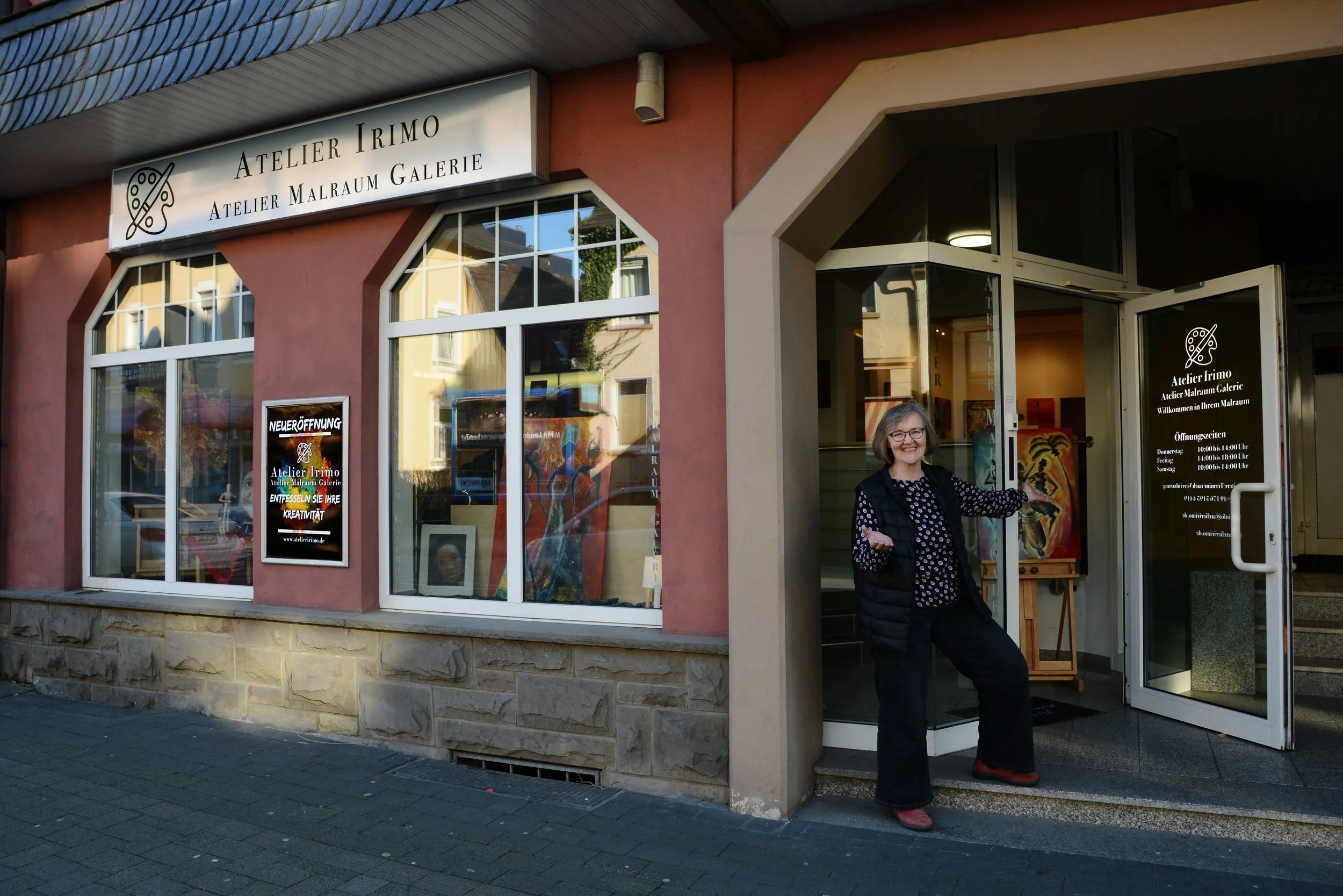 Das Bild zeigt die Eingangstür eines Kunstateliers mit einer freundlichen älteren Frau, die vor der Tür steht und sich freut, zur Galerie Am Eingang.