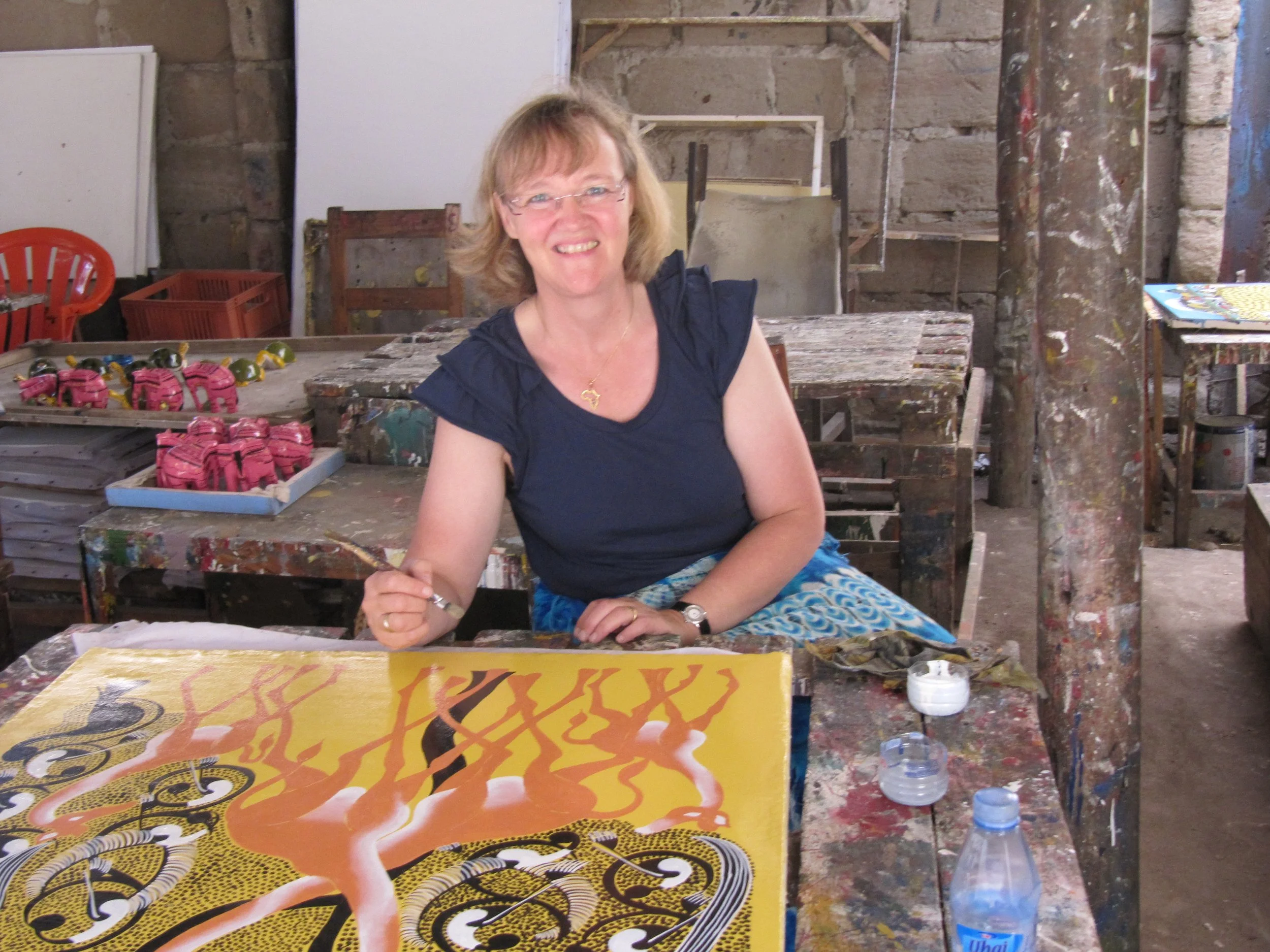 Eine lachende Frau sitzt in einem Kunststudio an einem Tisch, auf dem ein farbenfrohes, detailliertes Gemälde liegt. Im Hintergrund sind Regale mit vielen kleinen Kunstwerken und Farben.