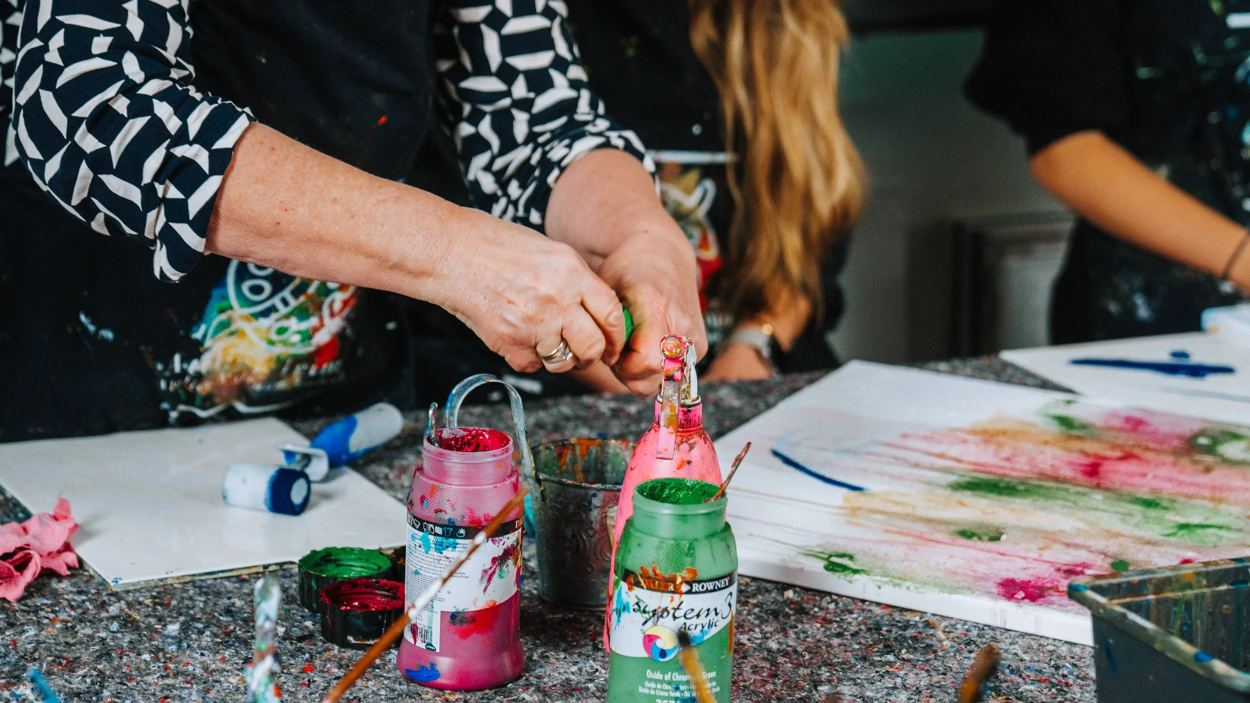 Menschen, die mit Fingerfarben auf Papier malen, auf einem Tisch mit Farbflaschen und Malutensilien.