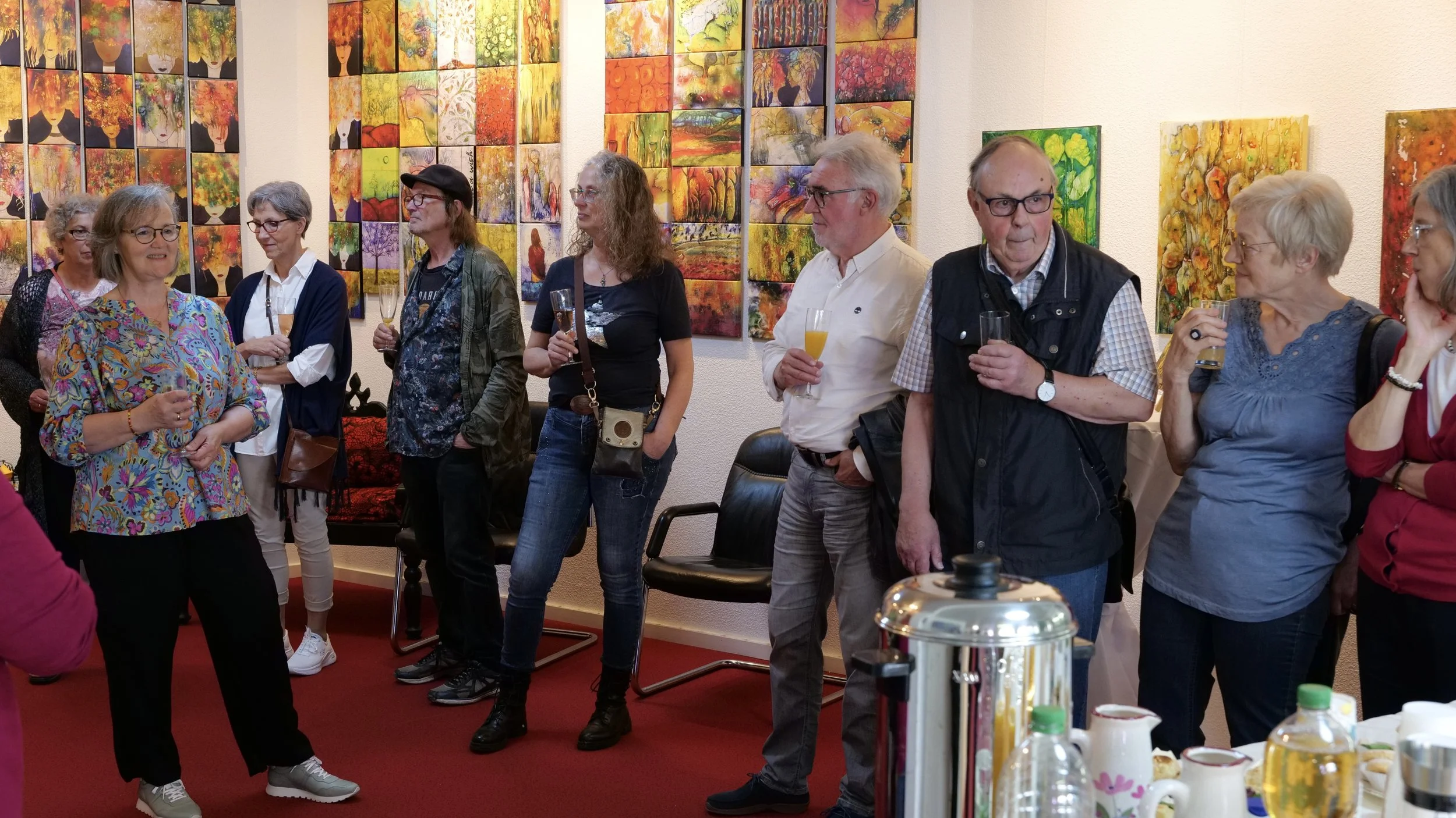 Menschen versammeln sich in einer Galerie bei einer Veranstaltung, im Hintergrund sind farbenfrohe Gemälde mit stilisierten Porträts von Frauen und Landschaften. Einige Personen trinken Getränke und unterhalten sich.