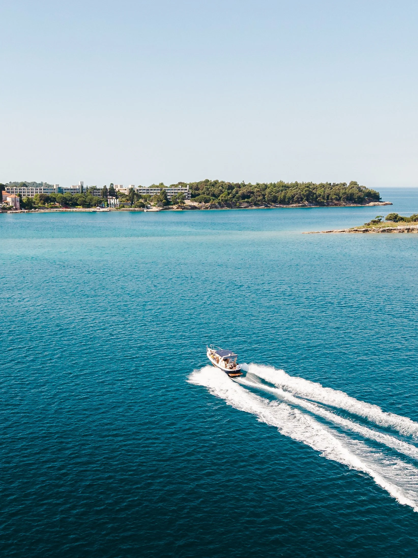 A motorboat speeding across turquoise water near a coastline with green trees and some buildings in the background.