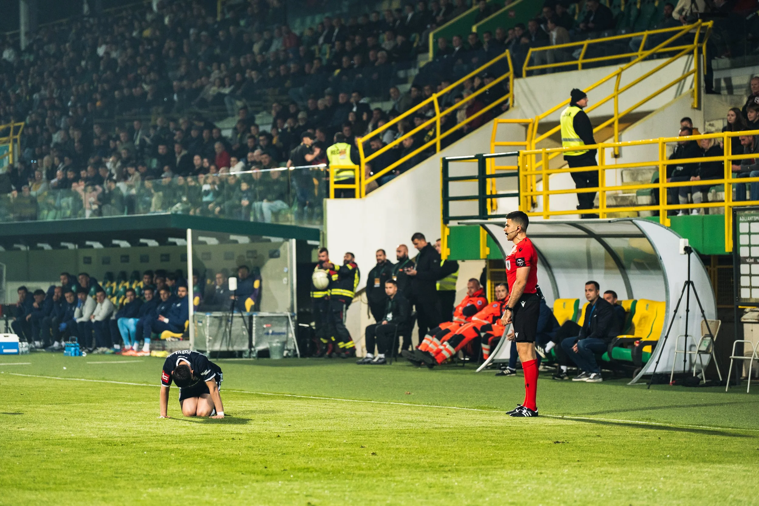 A soccer player kneeling on the field during a game with a referee standing nearby and spectators in the stands.