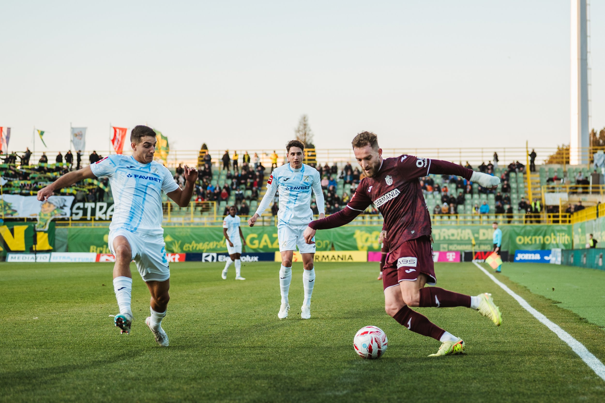 Soccer players in action on the field, with one player about to kick the ball while others move around him, and spectators in the background.