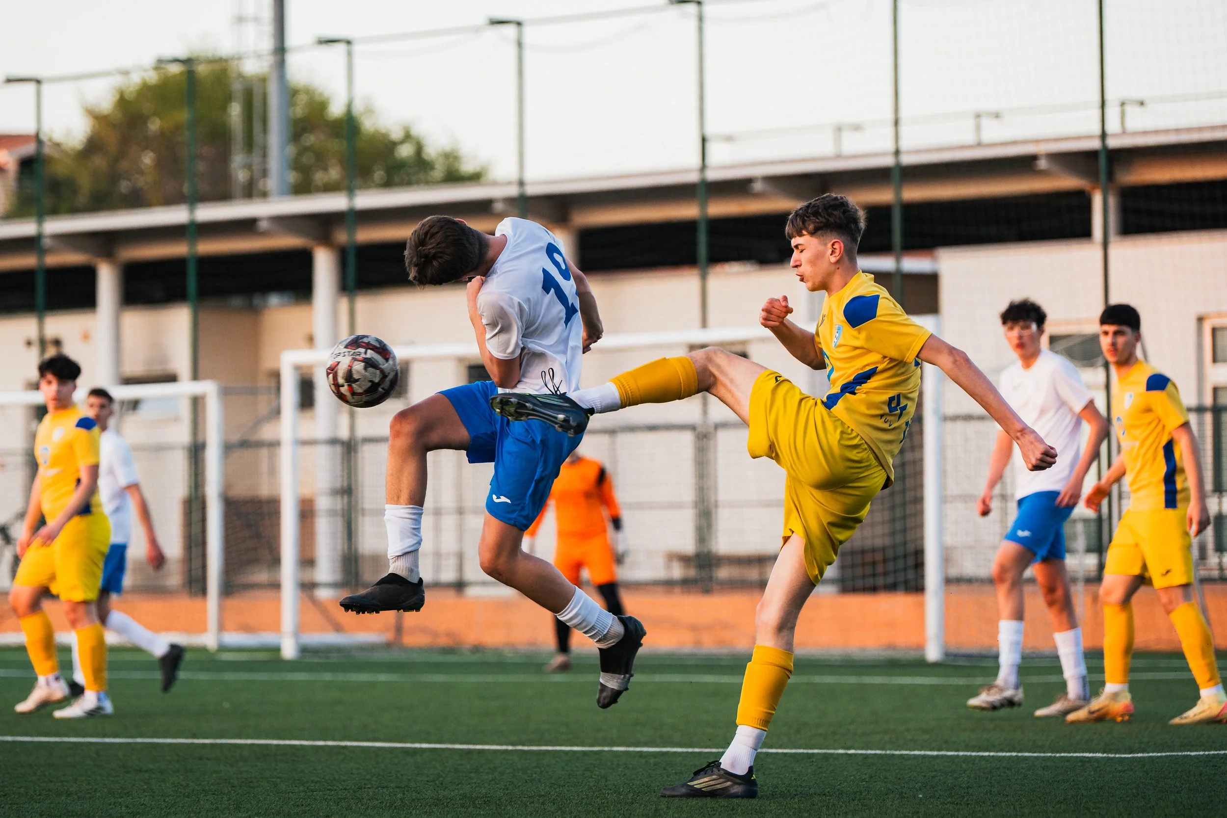 Two young male soccer players, one in a white and blue uniform and the other in a yellow and blue uniform, jump to contest a soccer ball during a game on a green field. Other players and a goalpost are visible in the background.