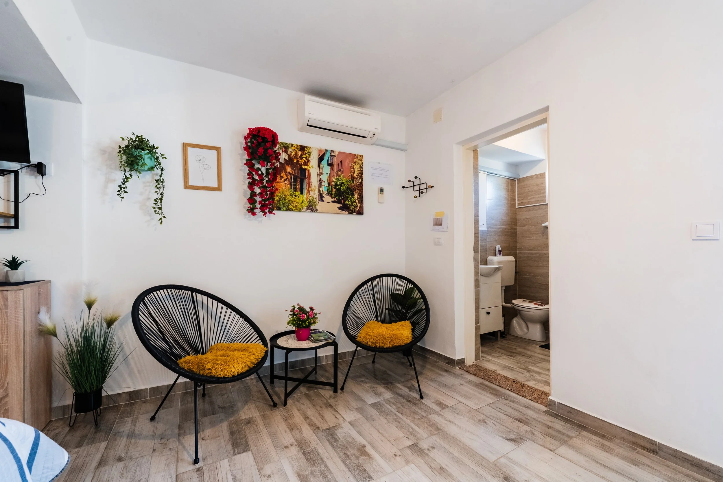 A cozy living room with two black wire chairs with yellow cushions, a small round table with a pink flower pot, and a variety of potted plants and wall decorations. There is a doorway leading to a bathroom visible in the background.