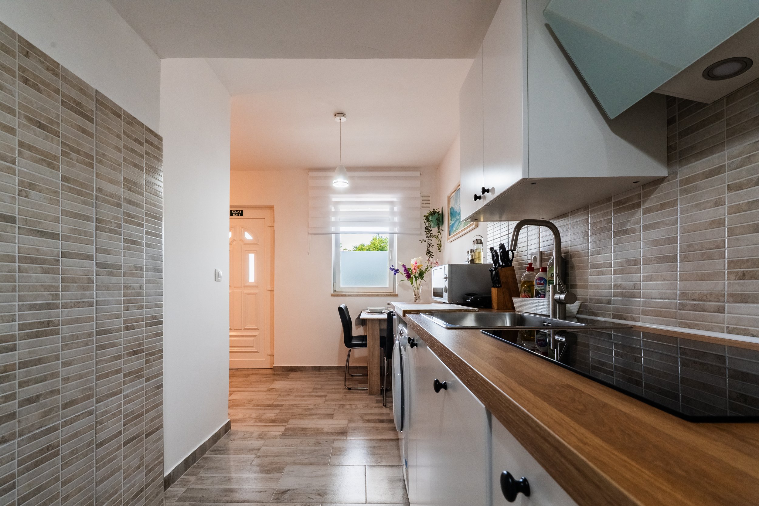 View of a kitchen with a beige tiled backsplash, wooden countertop, white cabinets, a sink, a stove, flowers in a vase, a microwave, and a window with blinds.