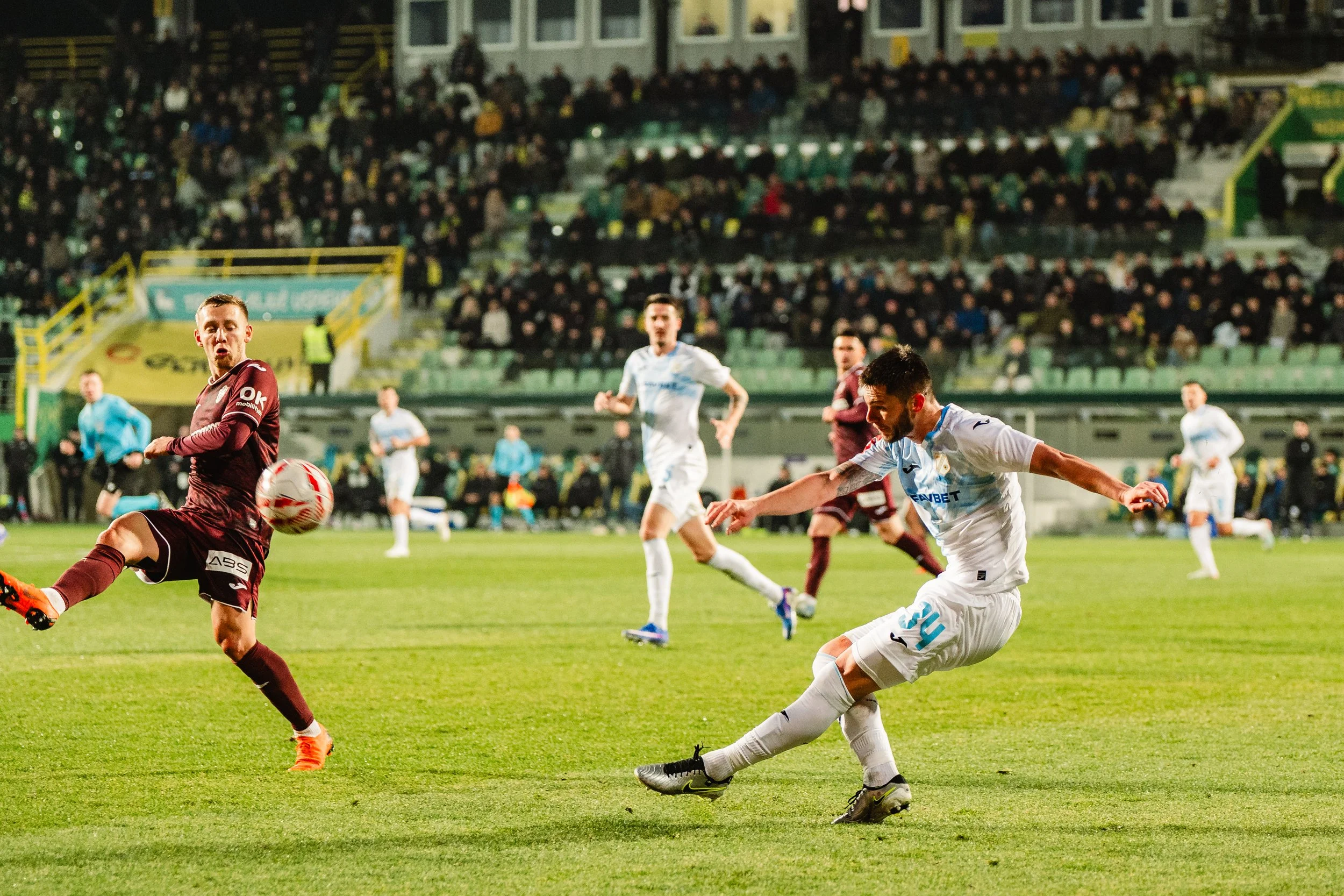 Soccer players compete for the ball during a match on a well-lit field in a stadium with many spectators.