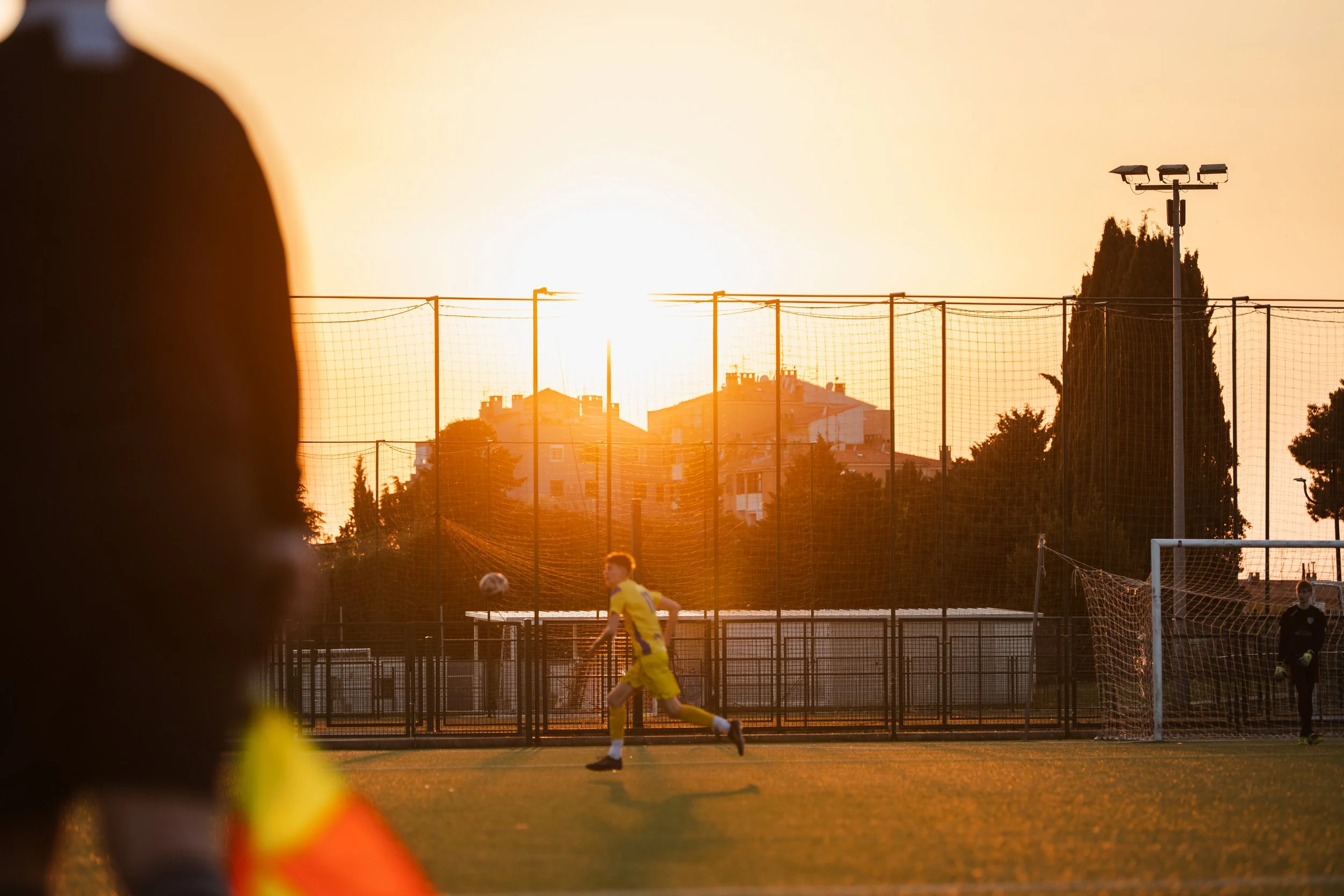 Children playing soccer on a field during sunset, with a foreground of an out-of-focus person wearing a sports jersey.