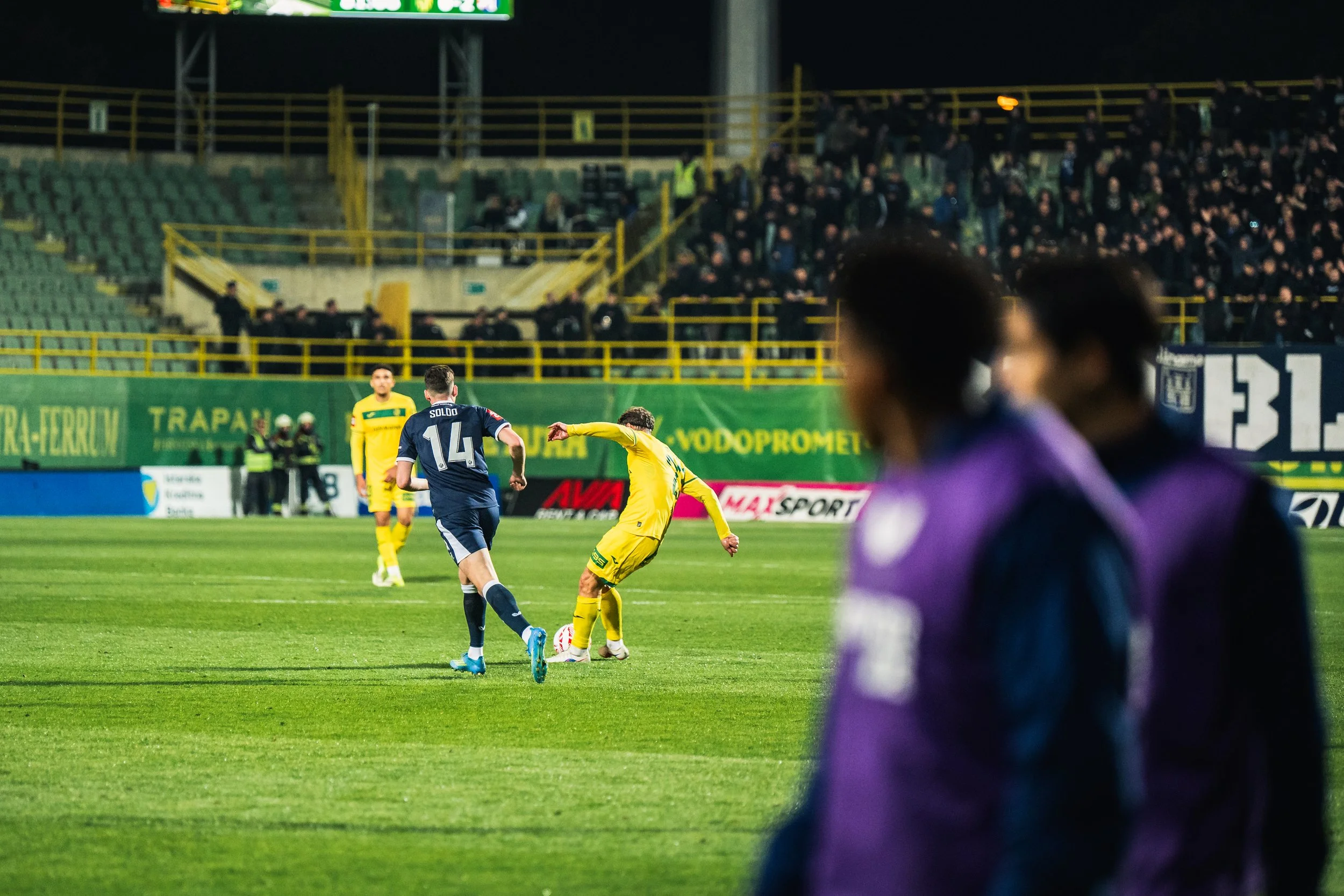 A nighttime soccer match with players in action on the field, crowds in the stands, and some spectators blurred in the foreground.