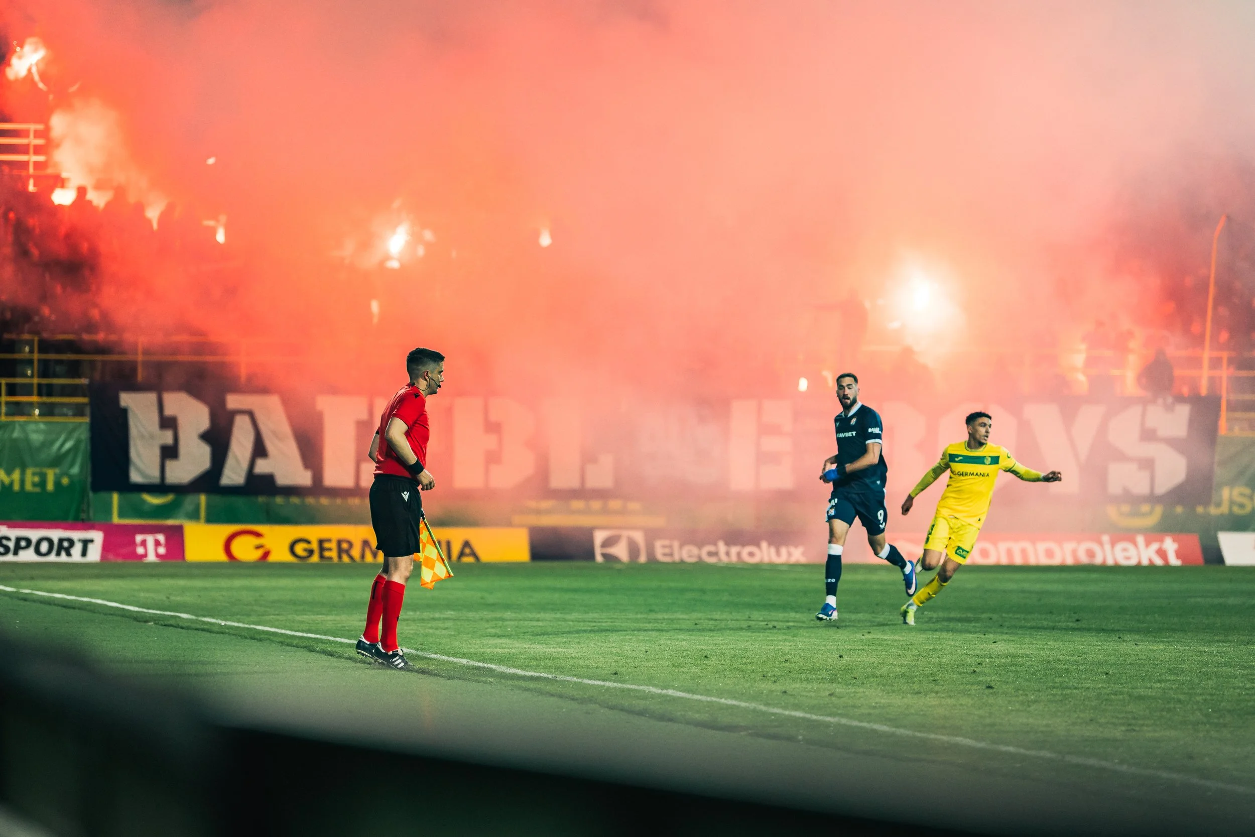 A soccer match at night with players on the field and large smoke or fire in the background emitting a bright orange glow, causing a smoky atmosphere.