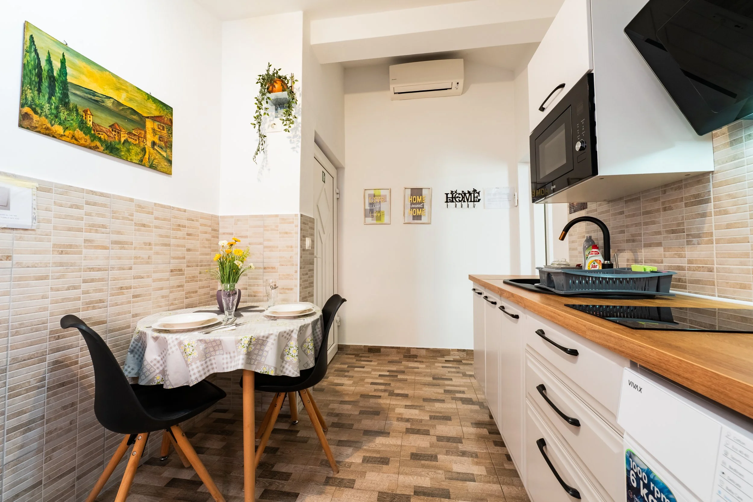 A dining table with a floral tablecloth, plates, glass, and purple vase with yellow flowers, situated next to a small kitchen area with white cabinets, black handles, a microwave, and a sink with a black faucet. The wall has framed decorative signs a