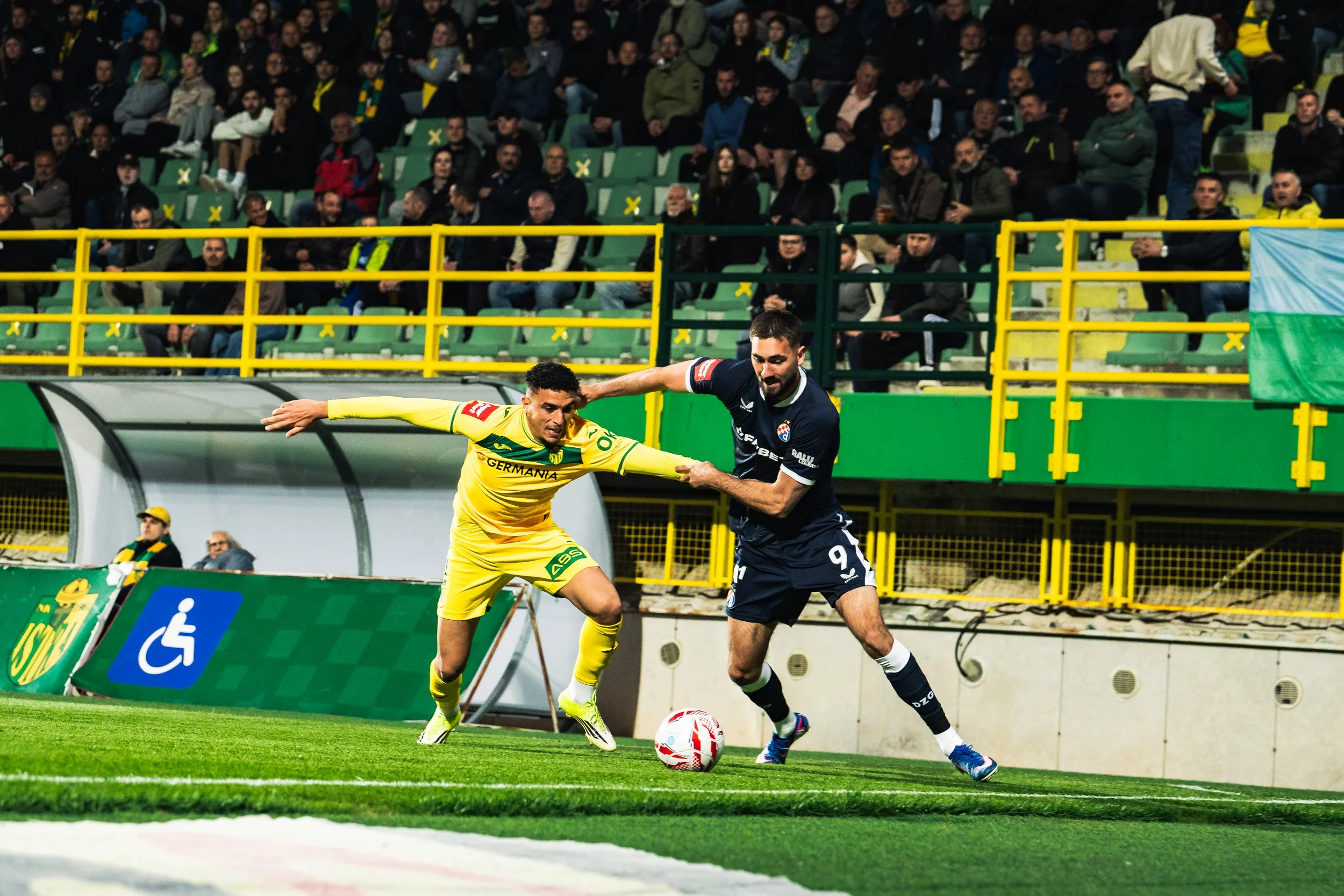 Two soccer players, one in a yellow jersey and the other in a navy jersey, competing for the ball on the field near the sideline, with a crowd of spectators in the background.