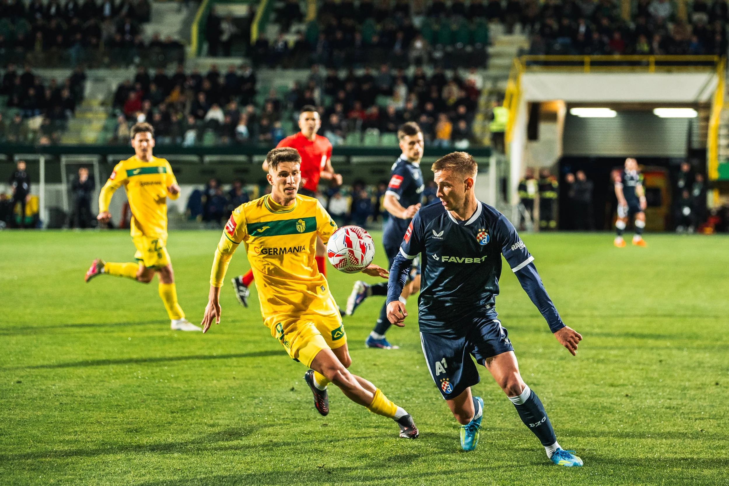 Soccer players in yellow and dark blue uniforms chasing a ball on a green field during a night game, with a crowd in the stands.
