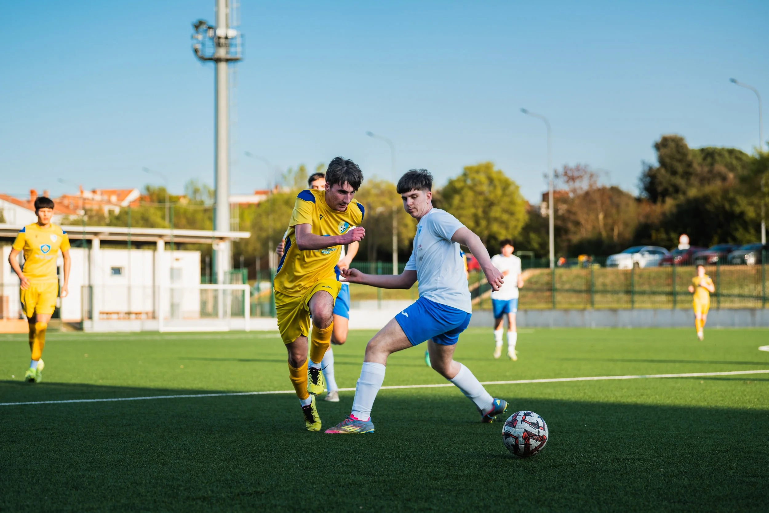 Two soccer players are competing for the ball on a green field with other players and a fence in the background.
