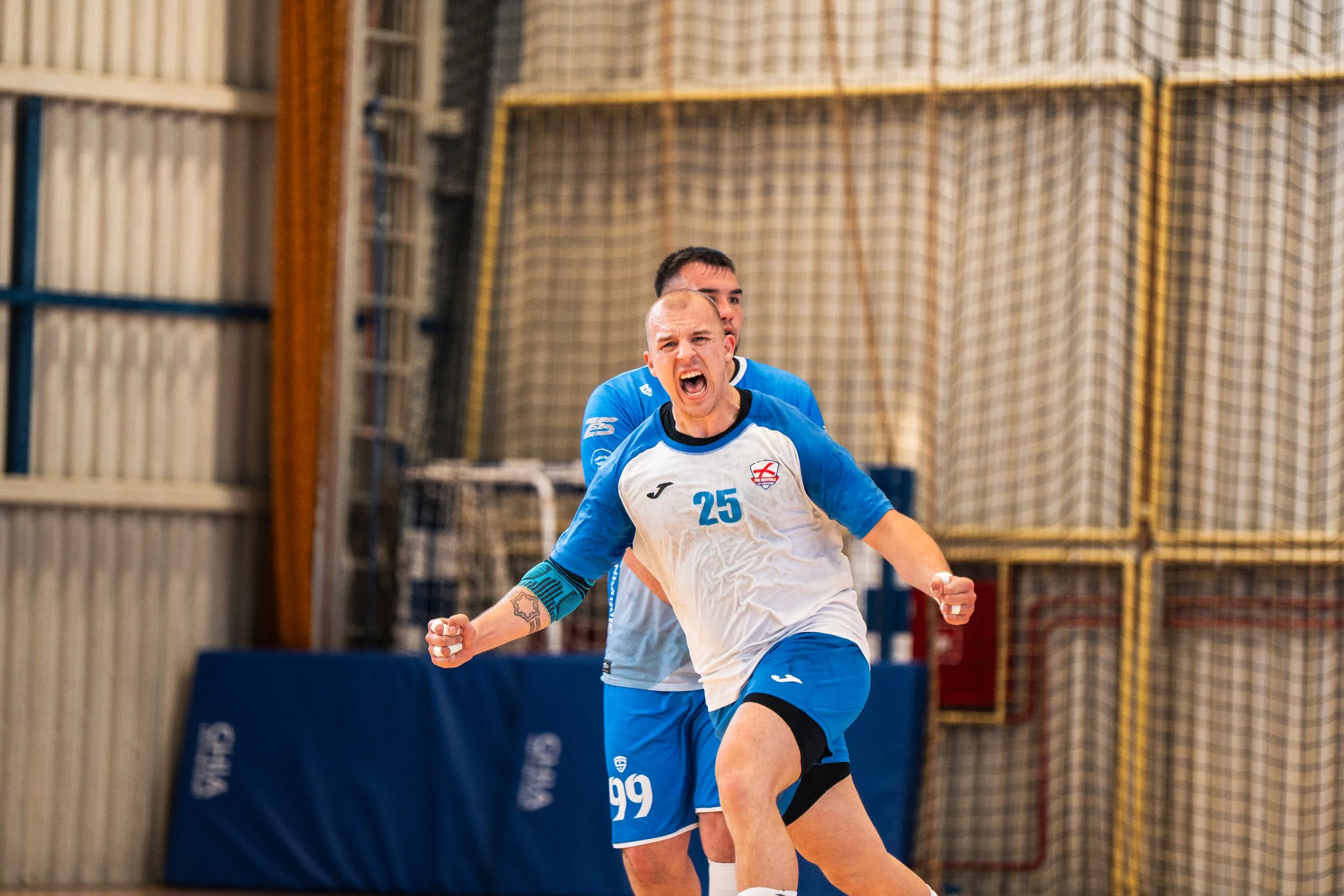 Two male athletes in blue and white jerseys celebrate on an indoor sports court, one with arms raised and clenched fists, in front of a net and sports equipment.