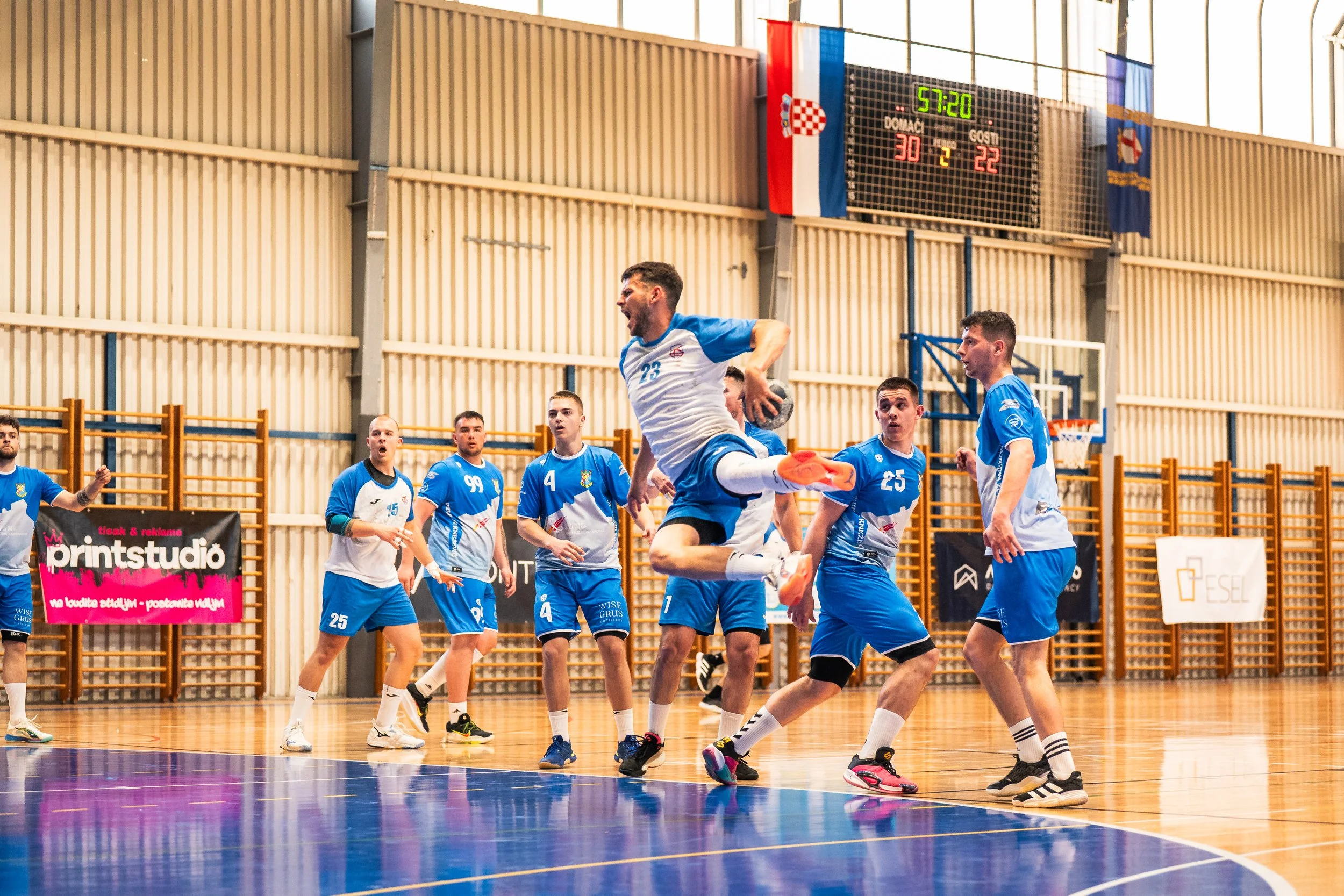 A group of male athletes playing handball in an indoor gymnasium. One player is mid-air heading towards the goal, while others watch or prepare. The gym has a wooden sports floor, flags hanging from the ceiling, and a scoreboard showing a score of 30