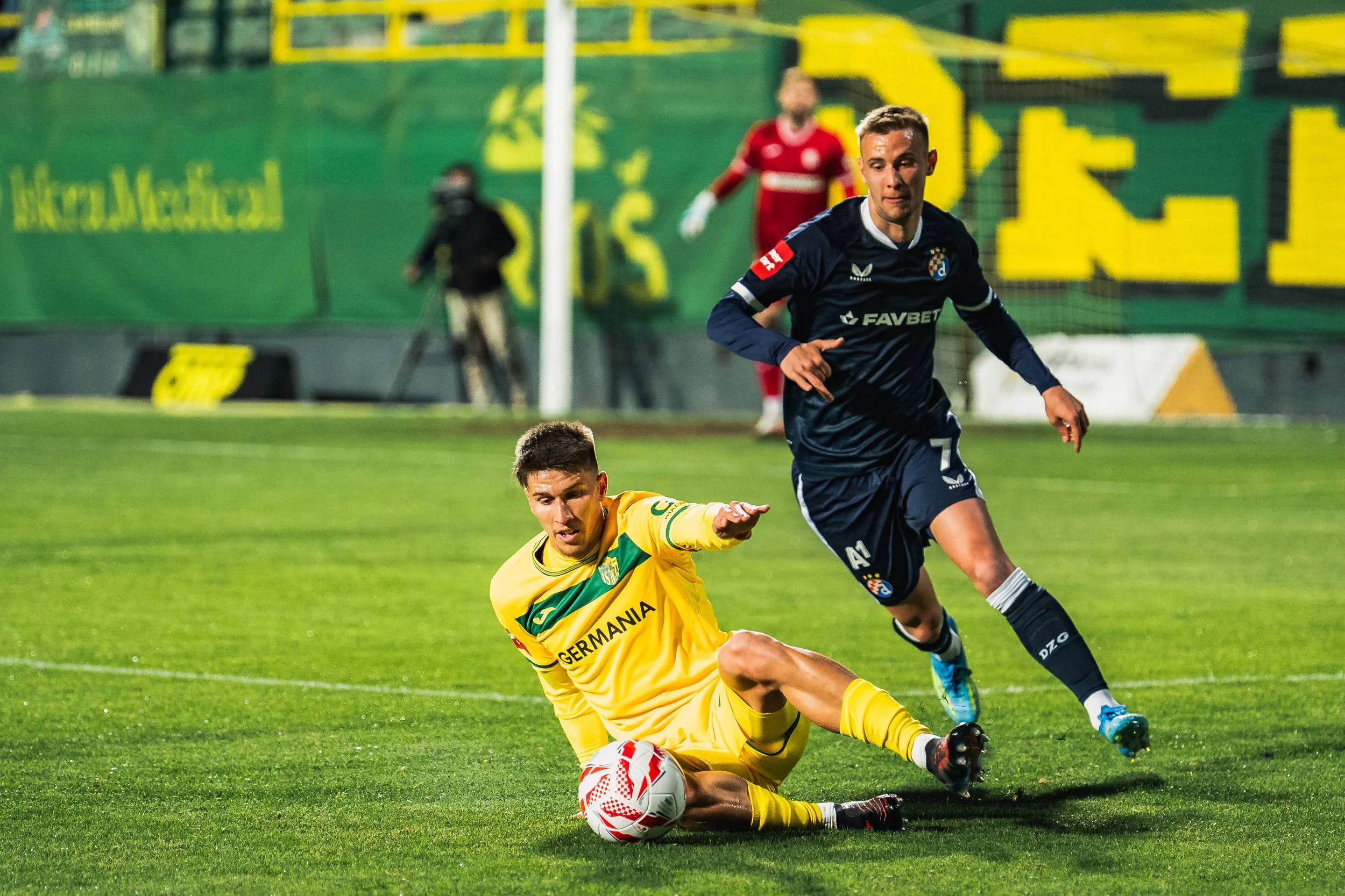 A soccer match showing a player from Germany in a yellow uniform on the ground reaching for the ball, while a player from Croatia in a dark uniform runs past him, and a goalkeeper in red is in the background.