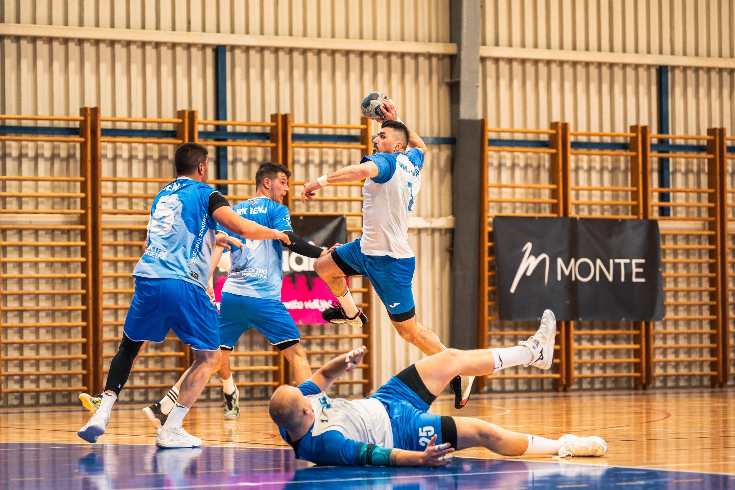 Handball game in progress; one player in white jumps with the ball, others in blue try to block or defend, with a player on the floor. Indoor gymnasium with wooden wall bars.