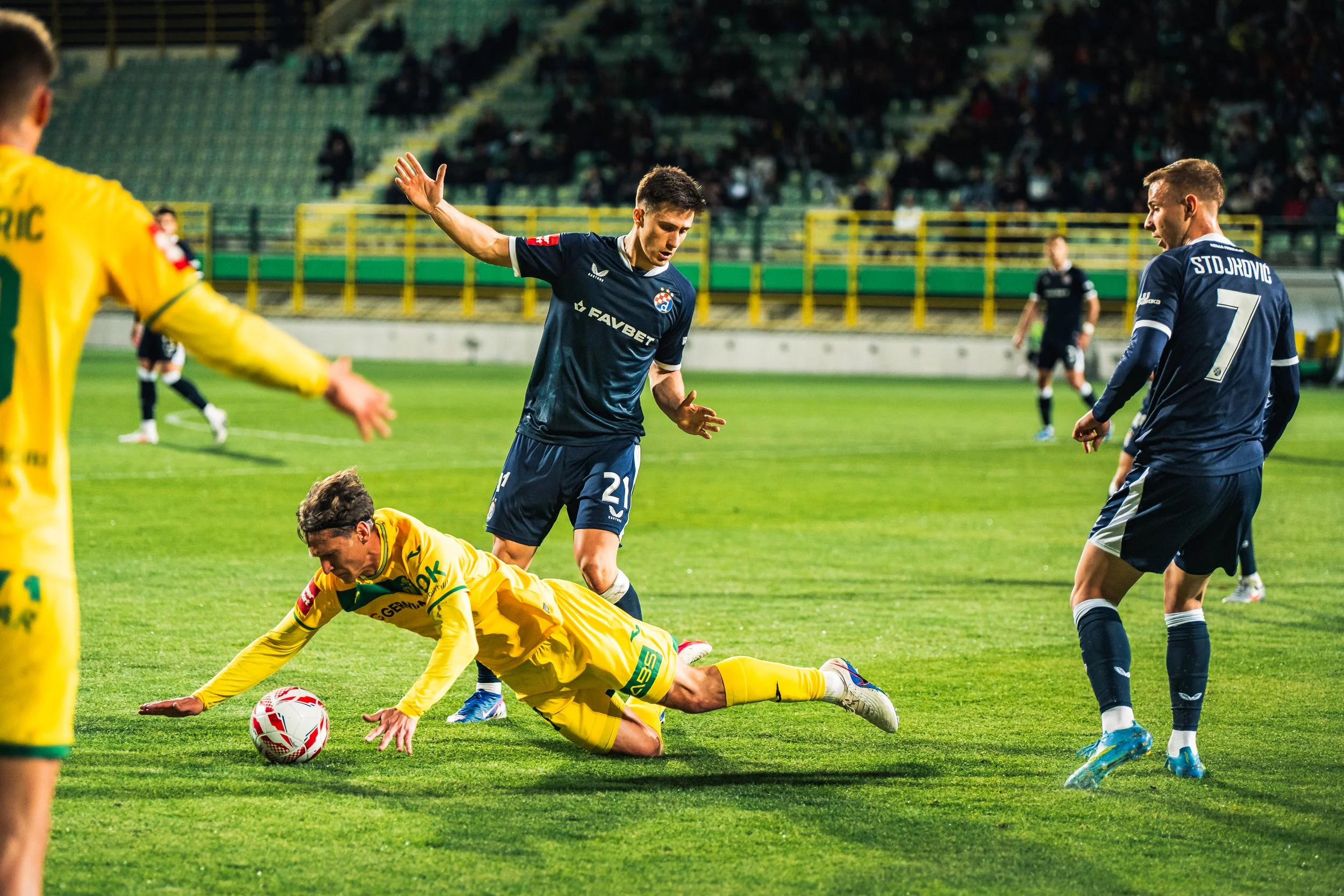 A soccer match with players in yellow and blue uniforms on a grass field. One player in yellow is on the ground reaching for the ball, while a player in blue is nearby. A referee or assistant in yellow is also visible.