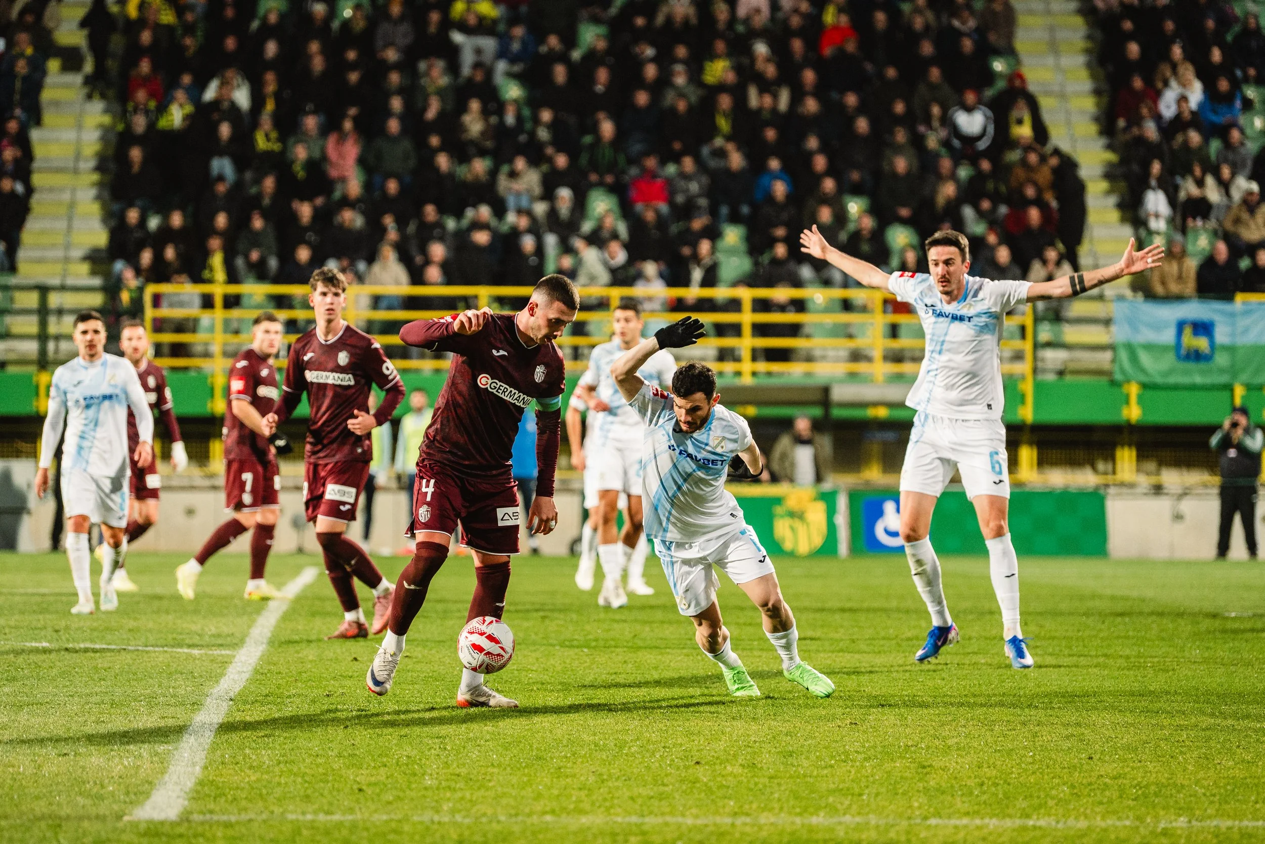 Soccer match with players in maroon and white jerseys competing for ball on field, with crowd in stadium background.