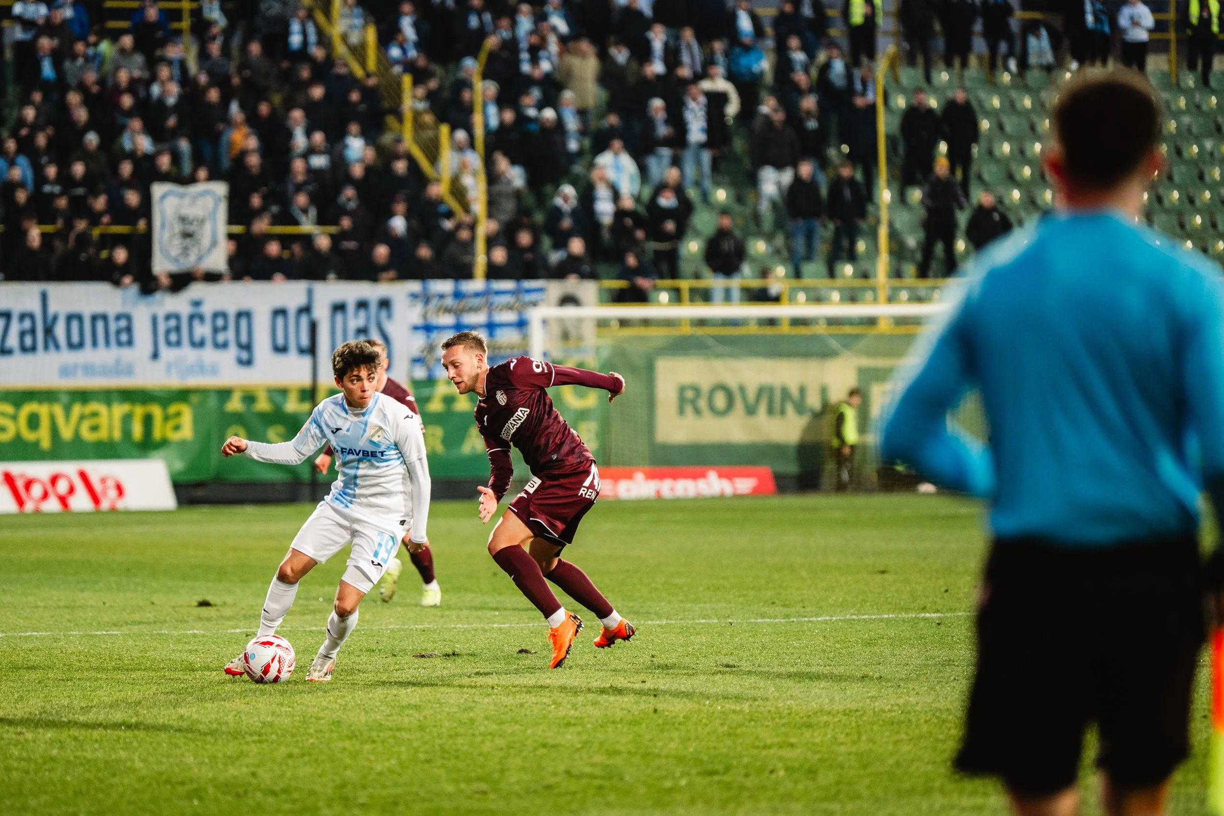 Two soccer players compete for the ball on a field, with a blurred crowd of spectators in the background and a person in a blue jersey in the foreground.