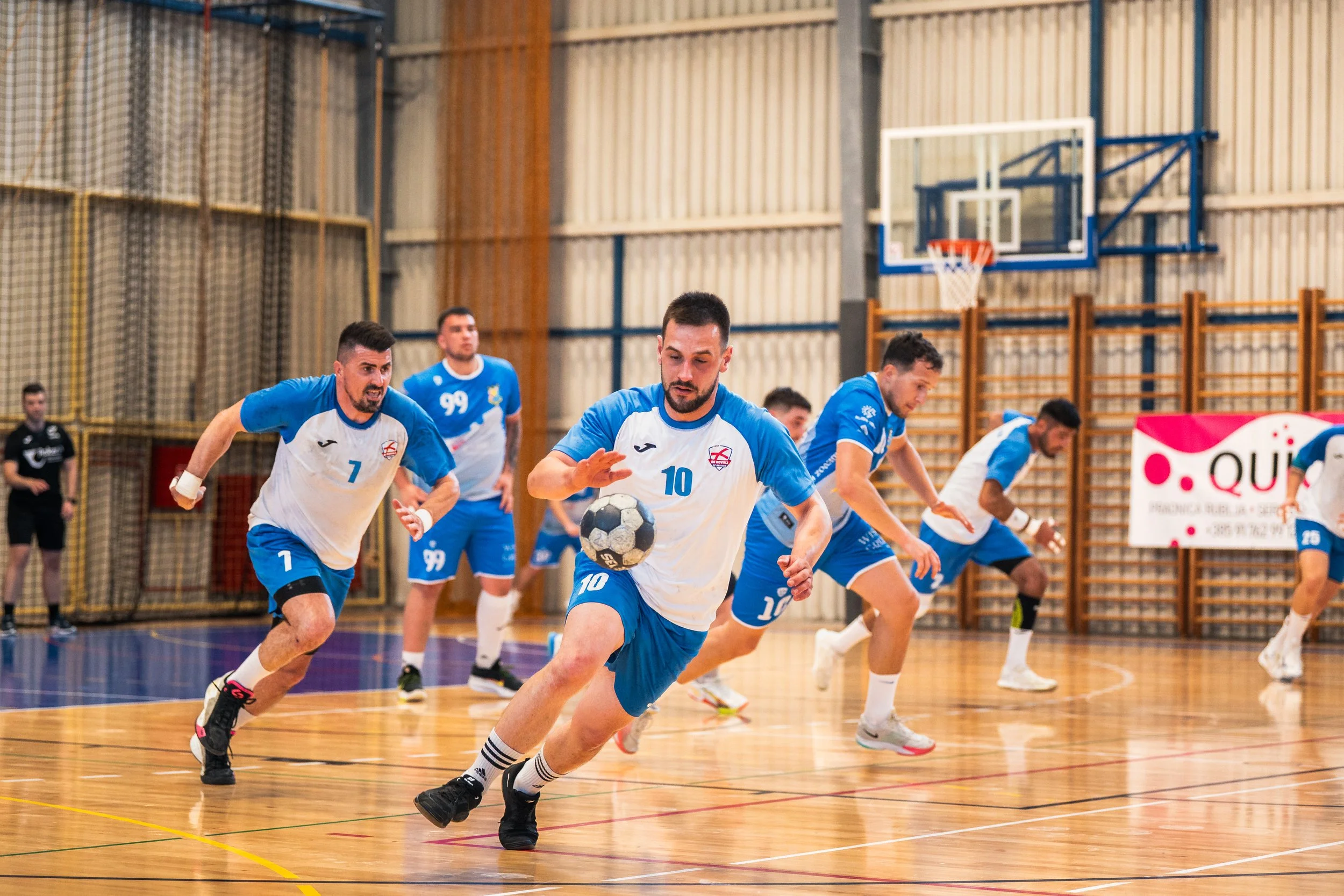 Indoor volleyball game with players in blue and white uniforms chasing the ball on a wooden court.