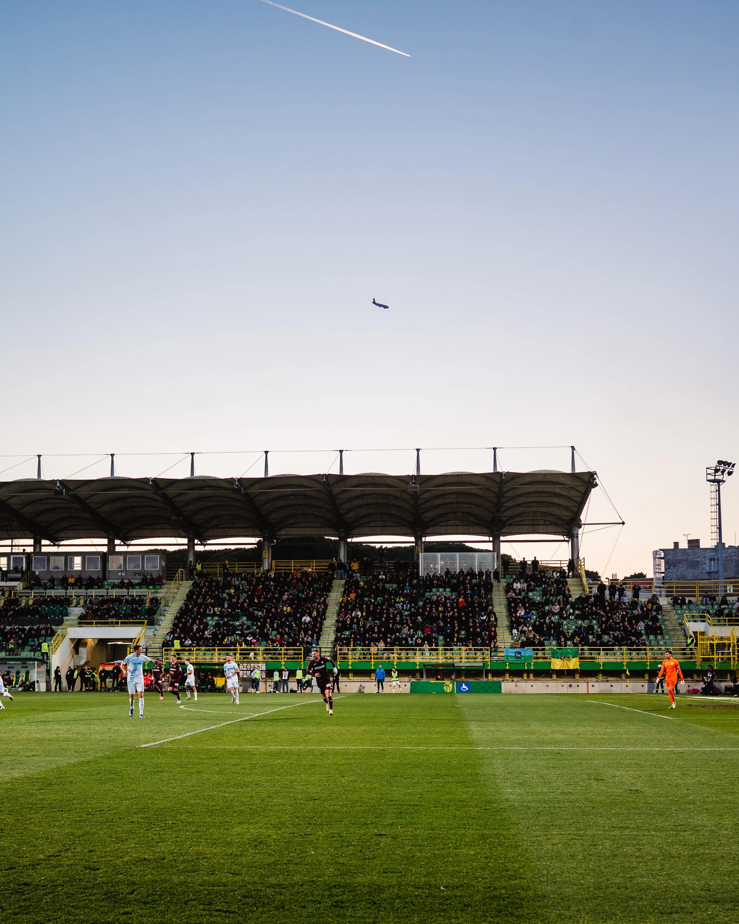 A soccer match in progress at a stadium. Players are on the field, with a group of spectators in the stands and a clear sky with a plane flying overhead in the background.