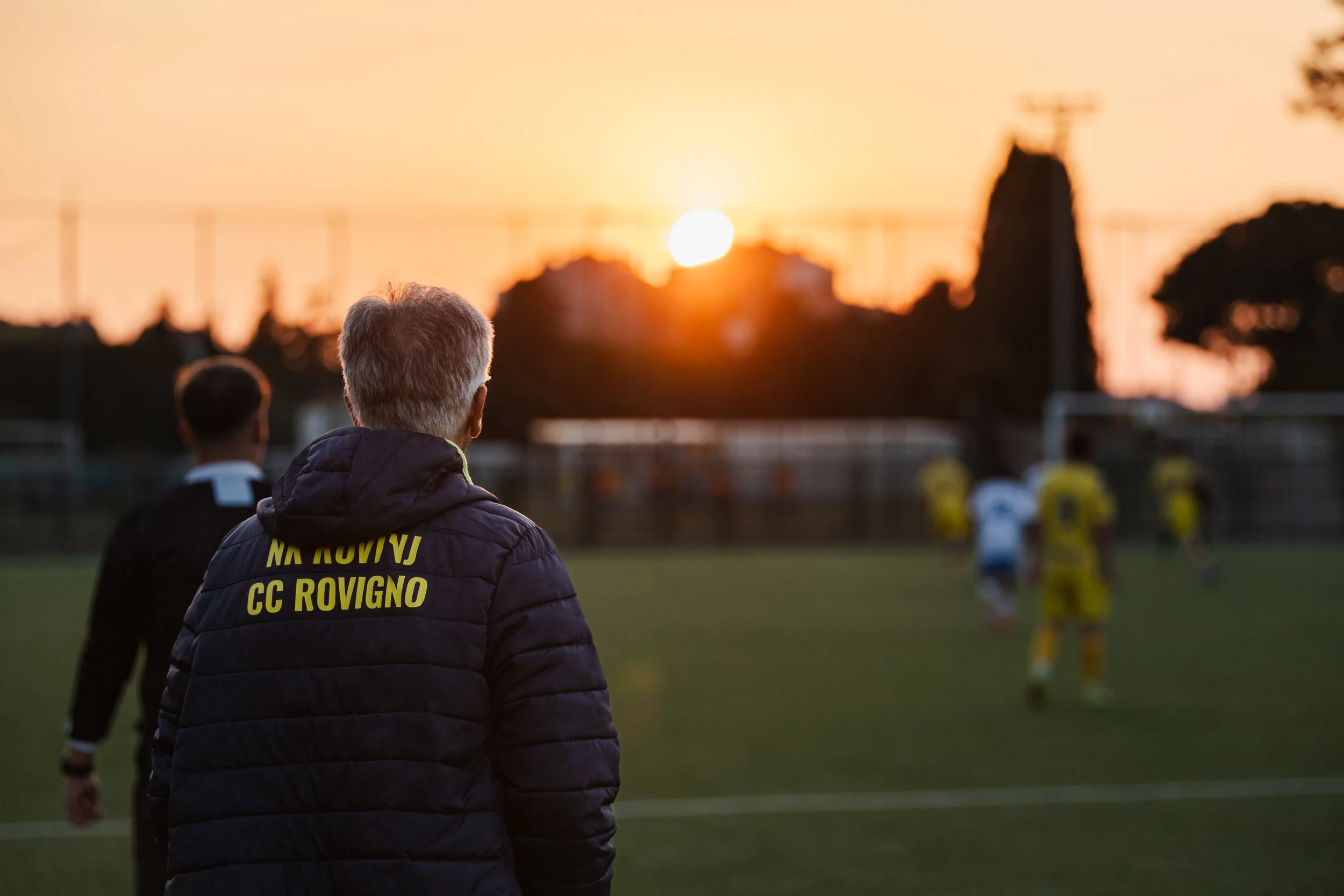 A person with gray hair wearing a black jacket with yellow text watching a sunset at a sports field during a soccer game, with other players in yellow uniforms in the background.