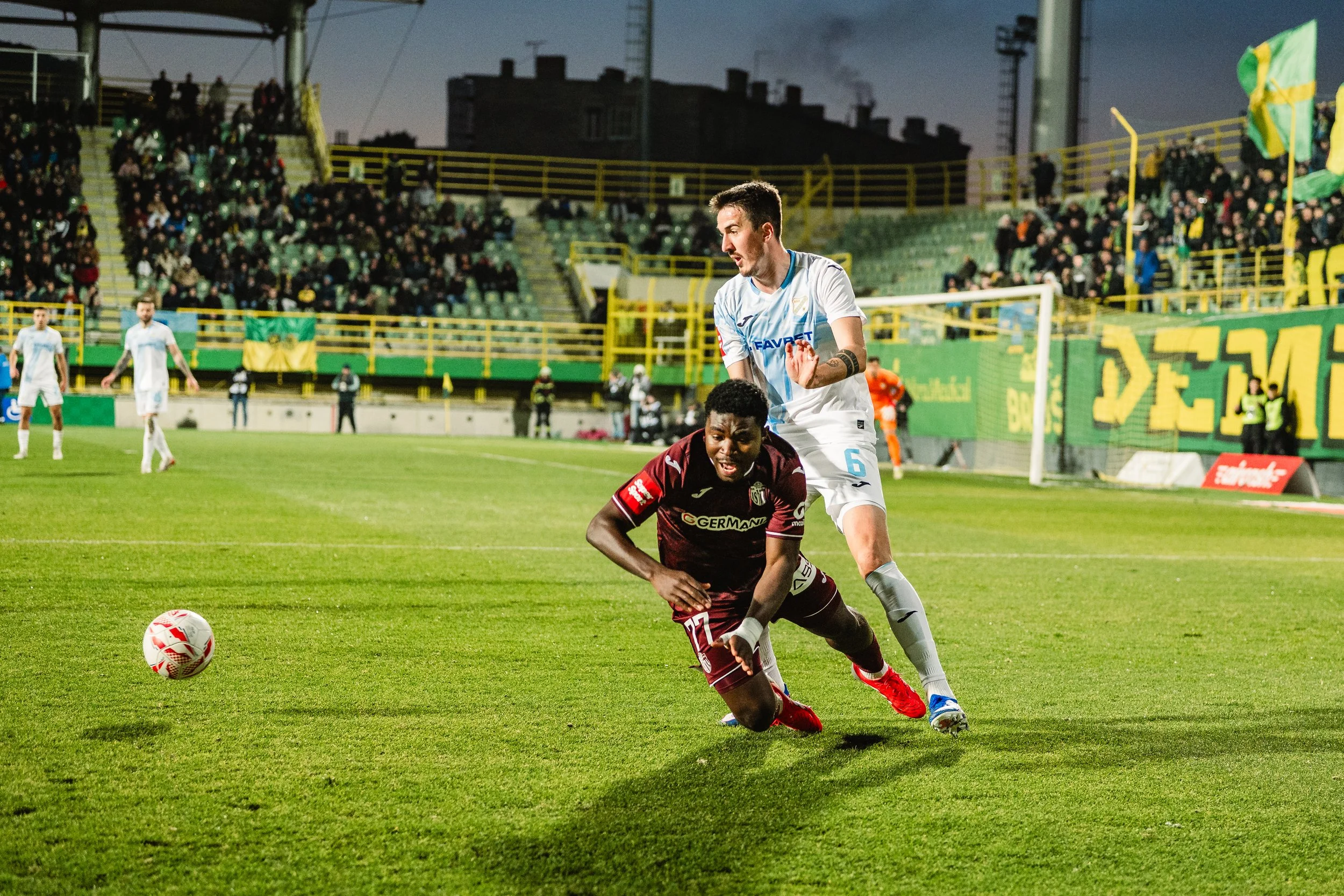 Two soccer players, one in a white jersey and the other in a maroon jersey, are competing for the ball on a soccer field during a game. The player in white appears to be reaching to block or defend, while the player in maroon is falling forward after