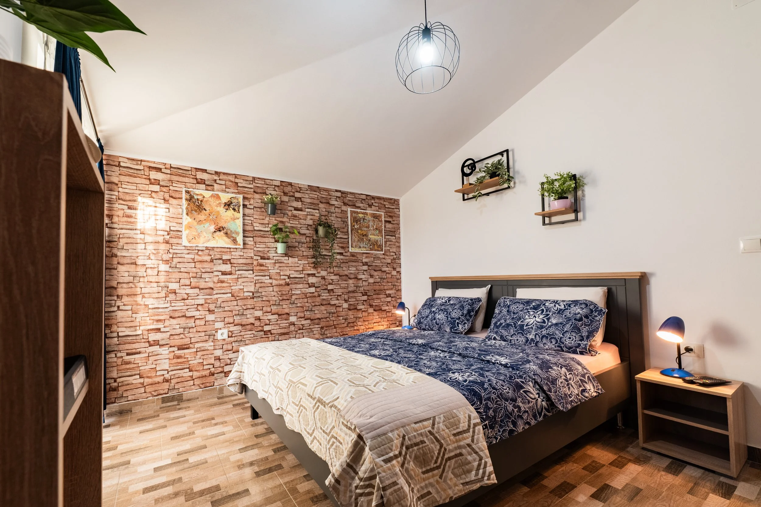 A cozy bedroom with a sloped white ceiling, a brick accent wall, and a bed with blue floral bedding. There are two small bedside tables with blue lamps, wall-mounted shelves with potted plants, and framed artwork on the brick wall.