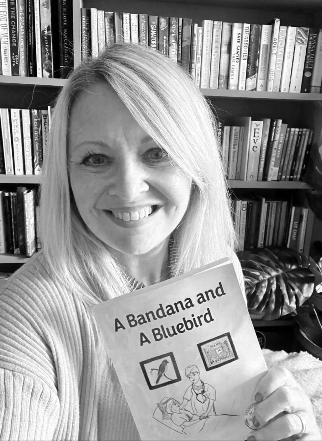 A woman with long blonde hair smiling and holding a children's book titled "A Bandana and A Bluebird" in front of a bookshelf filled with various books.