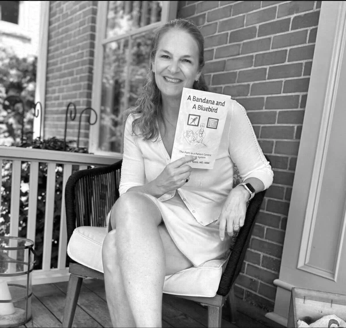 A woman sitting on a chair outdoors, holding a book titled "A Bandana and A Bluebird". She is smiling, wearing a white blouse and a watch, with a brick wall and a window in the background.