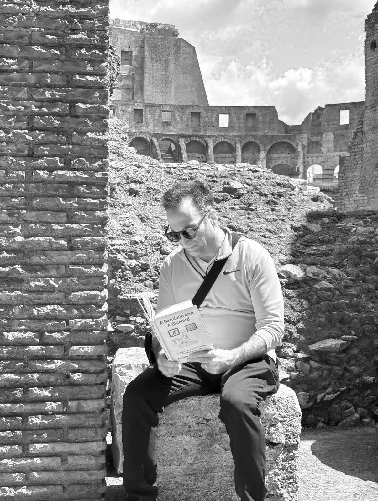 A man wearing sunglasses and a Nike sports shirt is sitting on a stone in front of ancient ruins, reading a book titled 'A Bandana and A Bluebird.' The ruins resemble the Colosseum in Rome, with arches and stone structures.