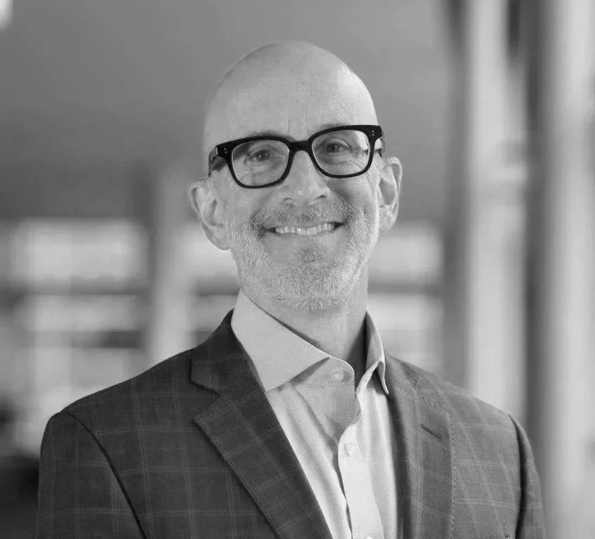 Black and white photo of a bald man with glasses smiling, wearing a suit in an indoor setting.