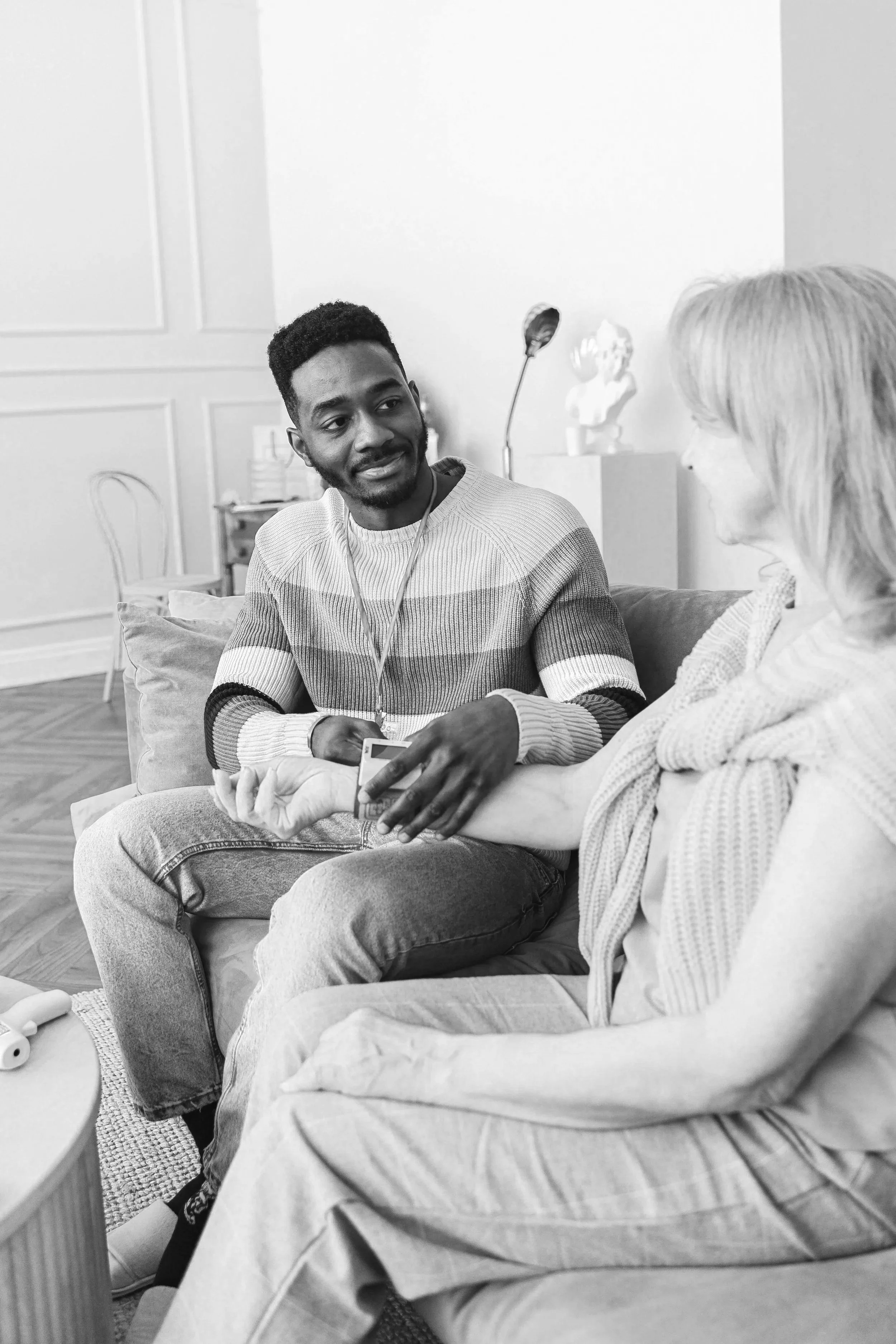 A young man and an older woman sit on a couch having a conversation in a living room. The young man is holding the woman's arm as she shows him her wrist.