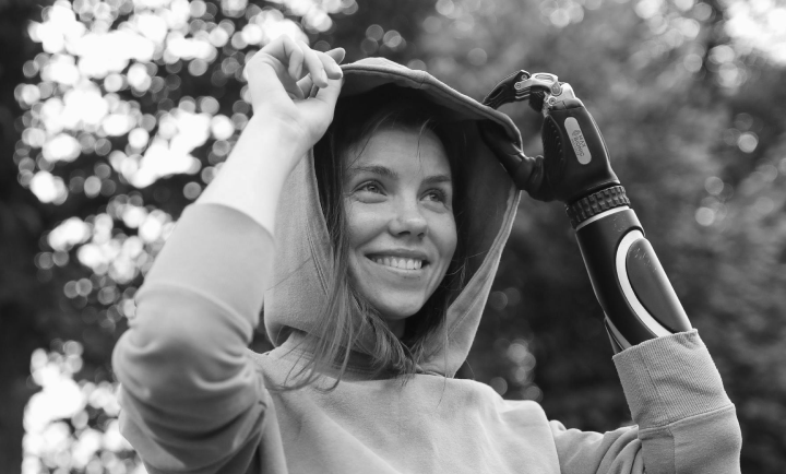 A woman outdoors pulling up the hood of her hoodie, smiling, with a background of trees and sunlight.