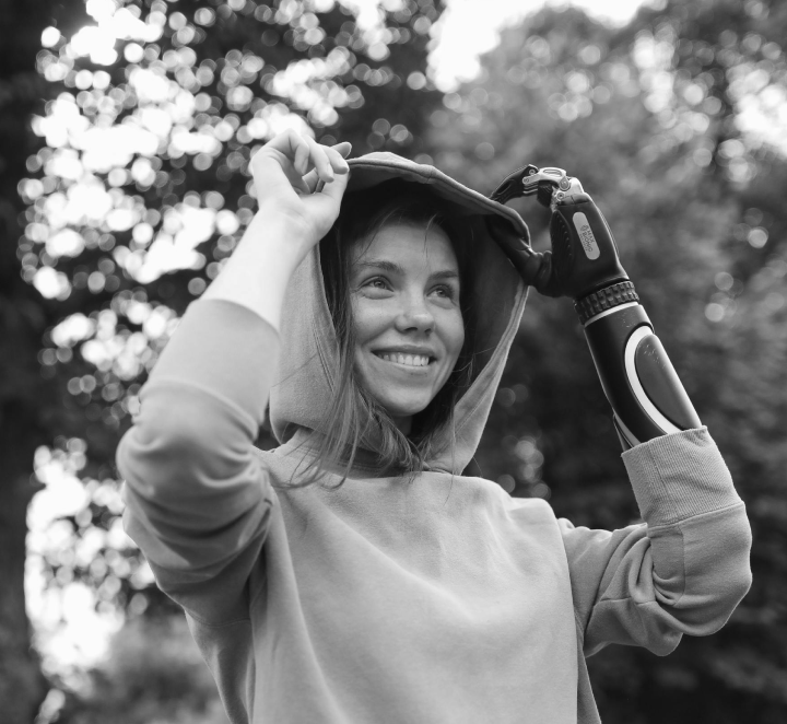 Young woman outdoors, smiling, wearing a hoodie, adjusting her hood, with an arm prosthetic, blurred trees in background.