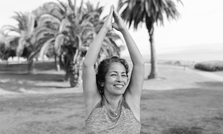 Woman practicing yoga outdoors near palm trees, smiling with hands in a prayer position above her head.