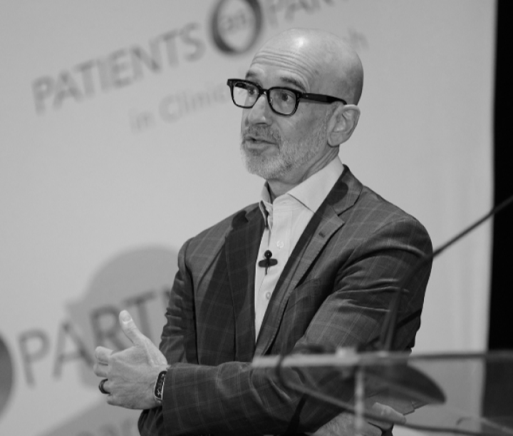 A man with glasses, a beard, and a bald head, dressed in a checkered blazer and white shirt, speaking at a conference or seminar.