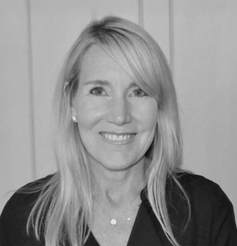 A smiling woman with long blonde hair, wearing a dark top and small earrings, standing in front of a wooden paneled wall.