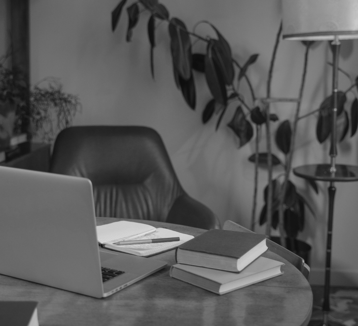 A modern office workspace with a laptop, open notebook, pen, and several stacked books on a round wooden table, with a black leather chair and a tall plant in the background.