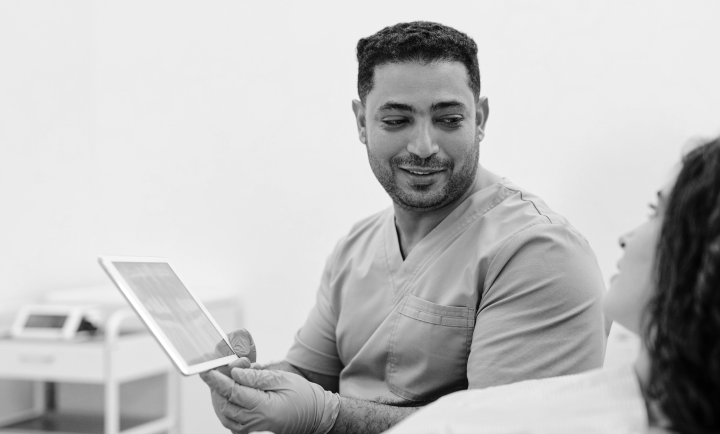 A male healthcare professional in scrubs holding a tablet and conversing with a patient in a medical setting.