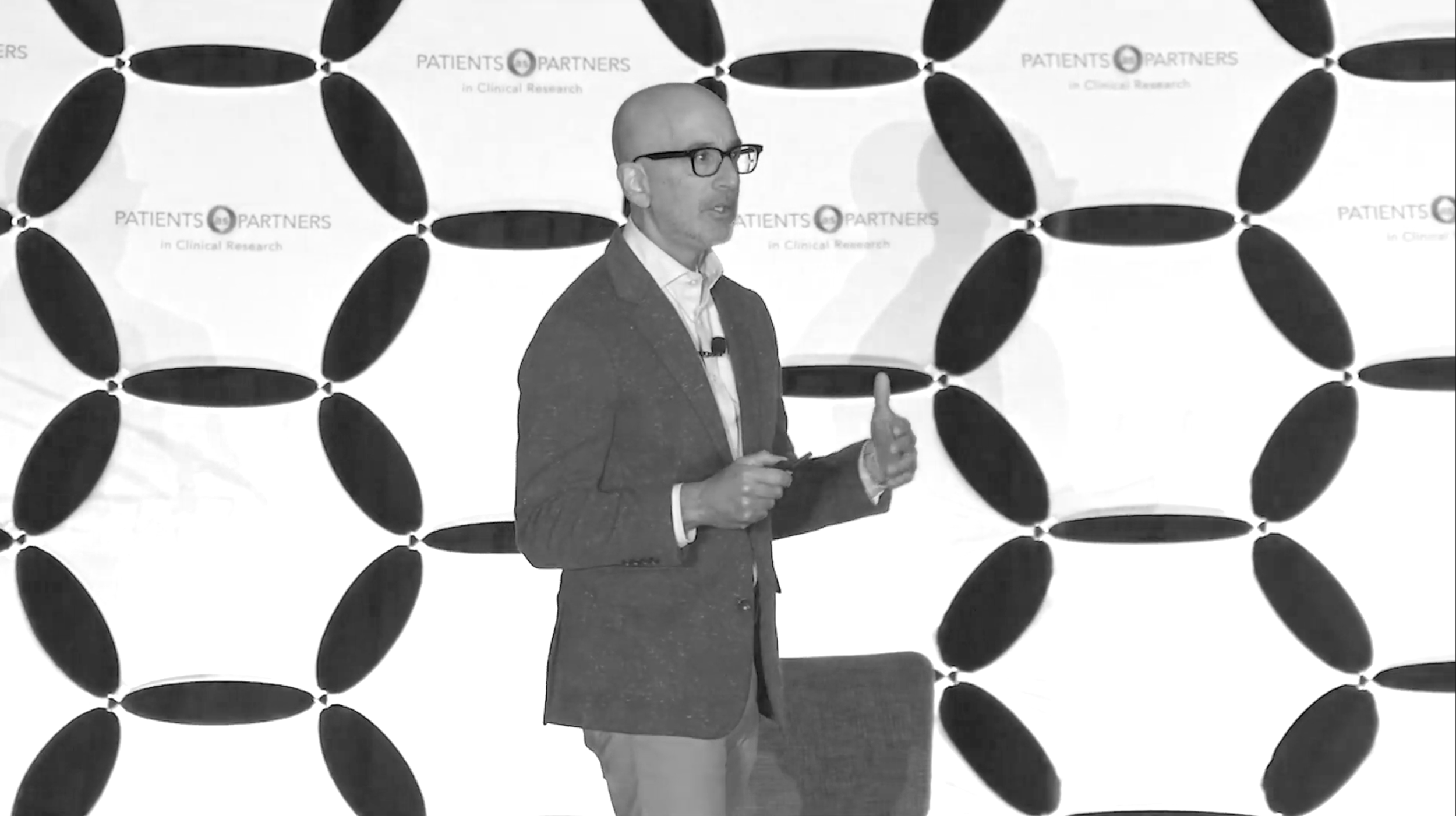 A man wearing glasses and a suit jacket giving a presentation with a backdrop that says "PATIENTS PARTNERS in Clinical Research."