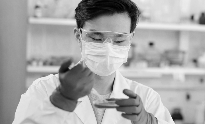 A scientist wearing safety goggles, a face mask, and gloves examining a petri dish in a laboratory.