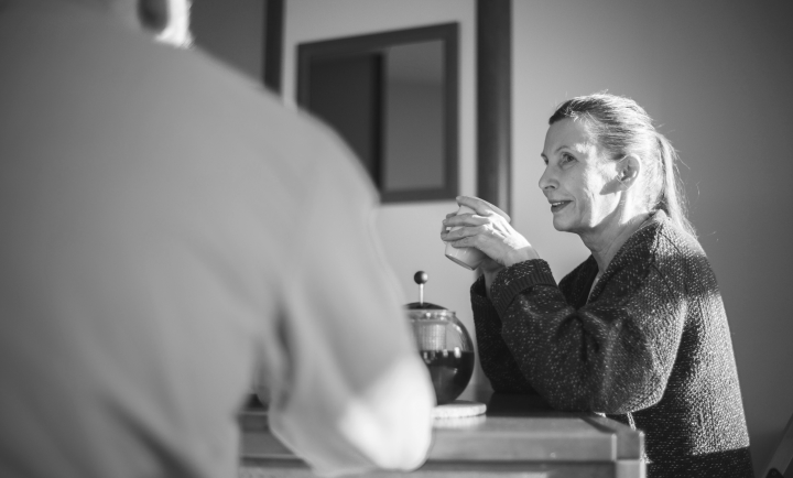 A woman sitting at a table, holding a cup, engaged in conversation with another person whose back is to the camera. The woman is smiling and wearing a dark, textured sweater.